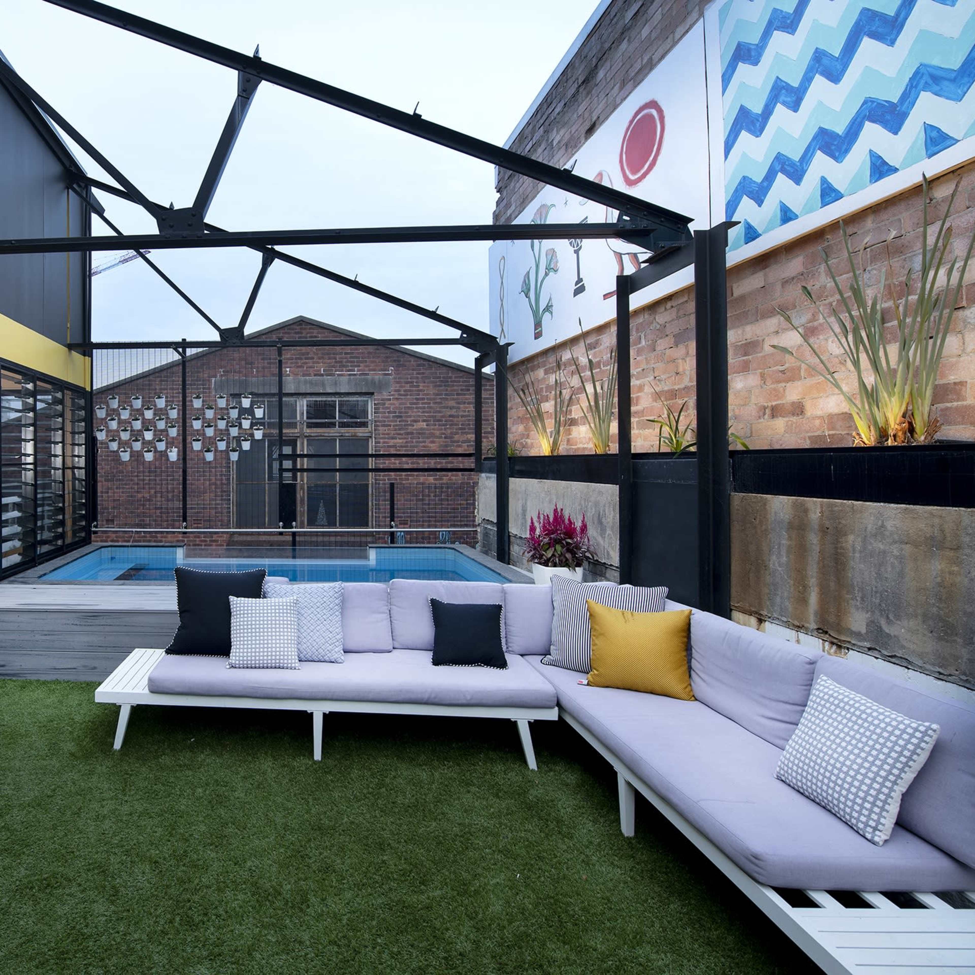 A modern outdoor lounge area with a swimming pool, featuring a sectional sofa with yellow and gray cushions and a backdrop of a brick wall and colorful murals.