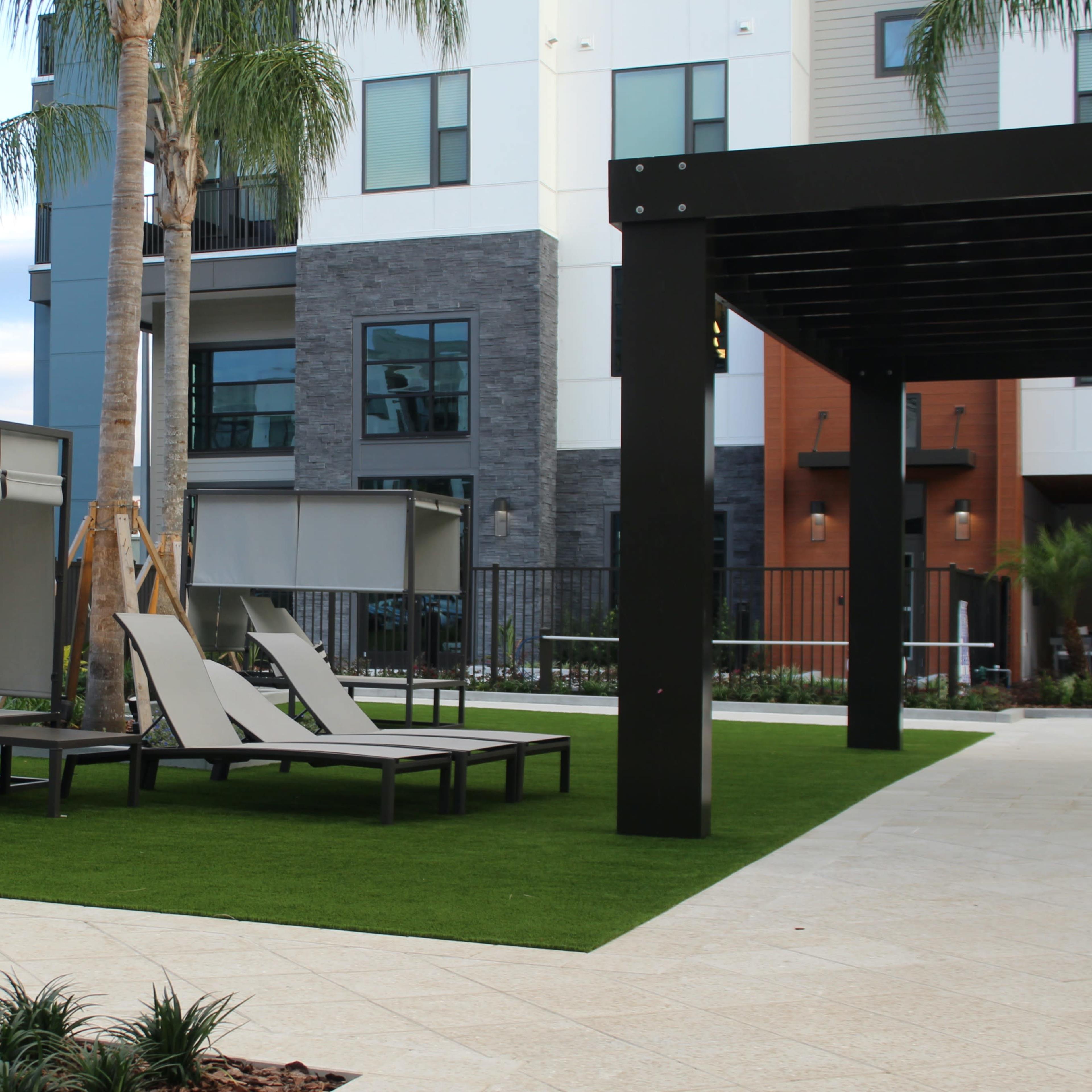 A landscaped outdoor area with lounge chairs, a shaded seating structure, and green artificial grass near an apartment building.