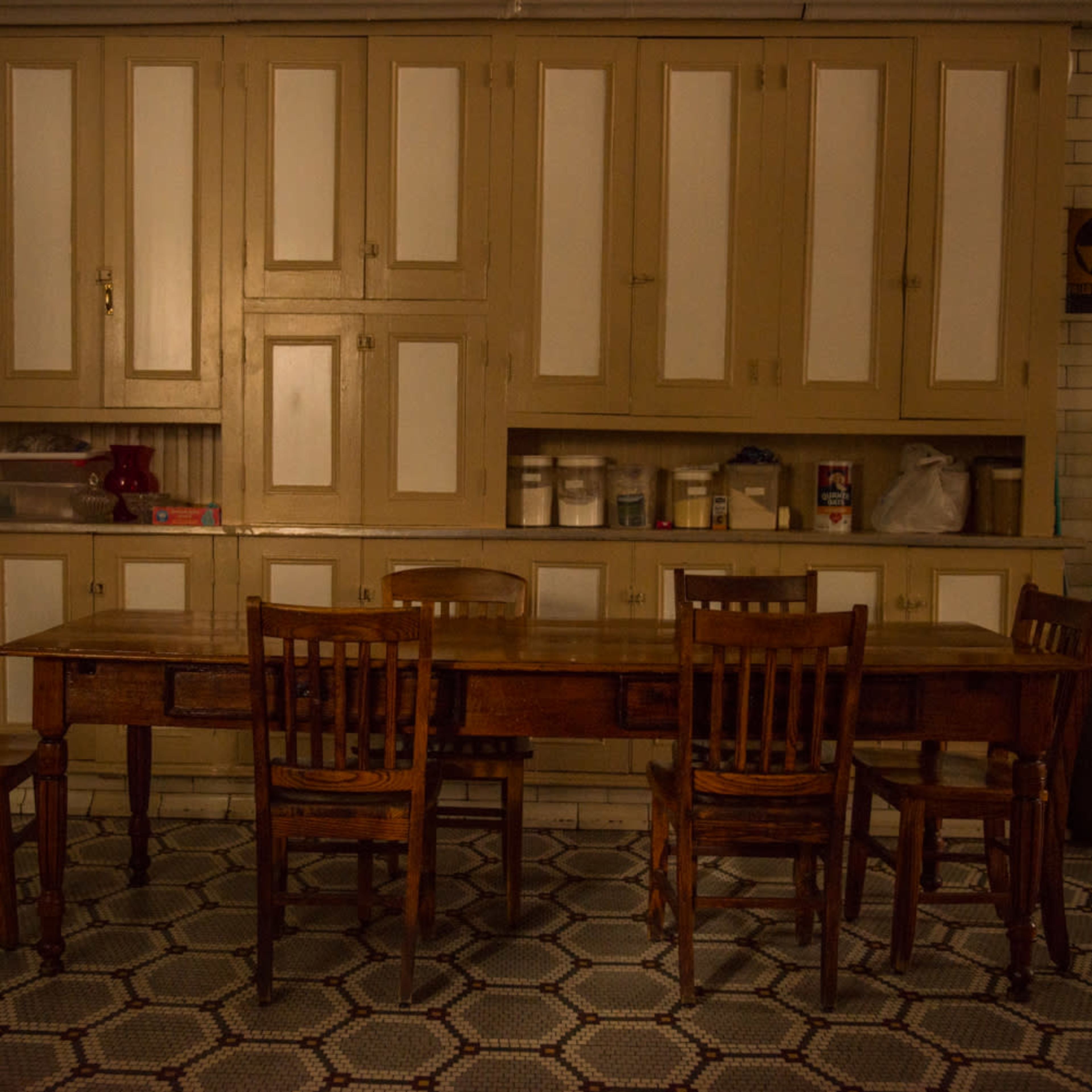 A long wooden table surrounded by four wooden chairs sits in a dimly lit room with a cabinetry wall in the background.