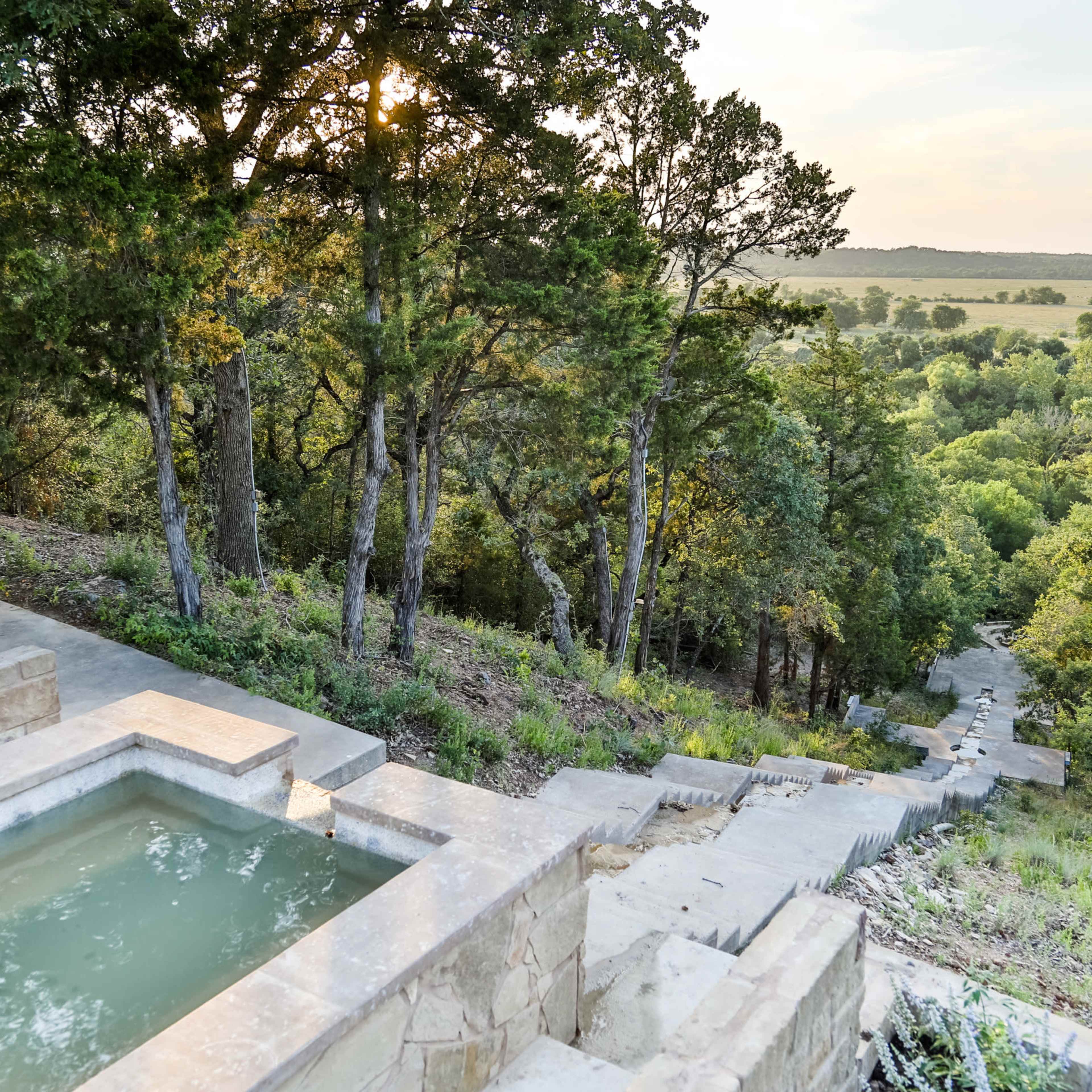 A stone staircase leads down from a small pool surrounded by trees, descending towards a green valley beyond.
