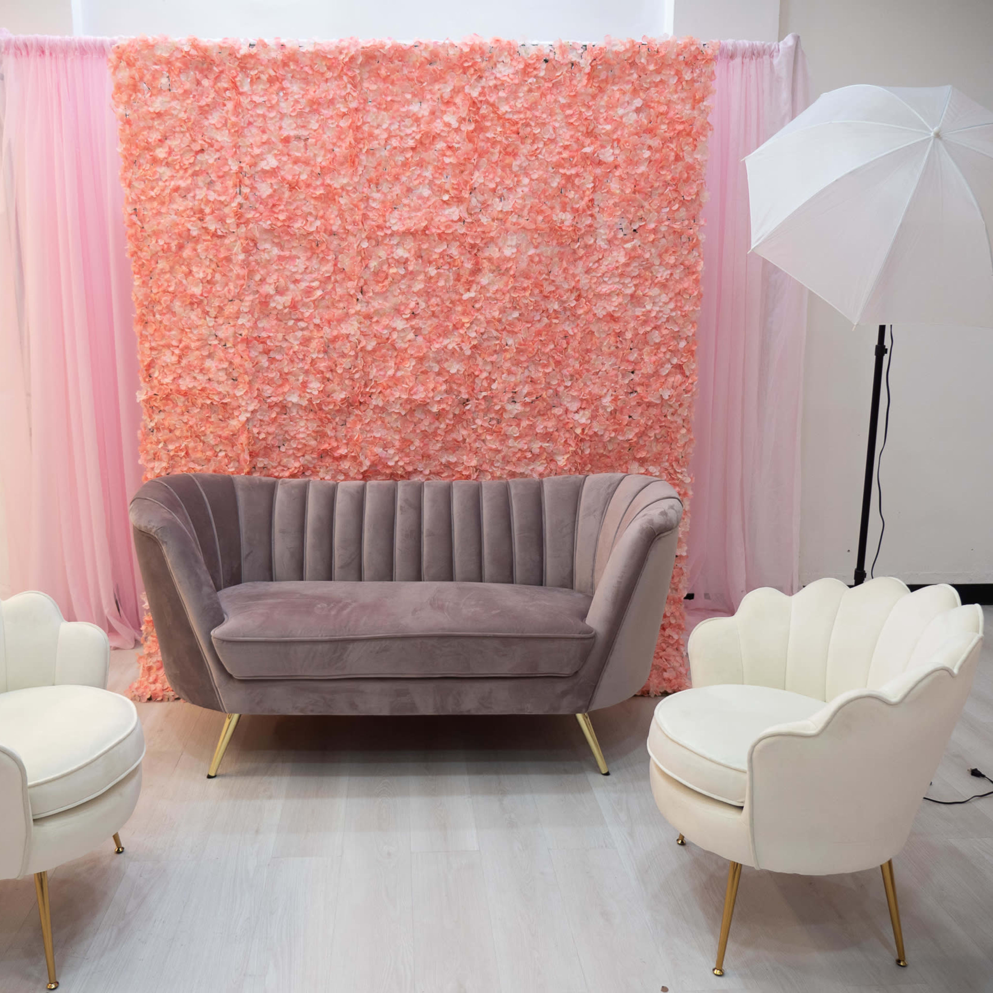 A stylish seating area with a grey velvet couch and two white chairs, set against a backdrop of pink floral decorations.