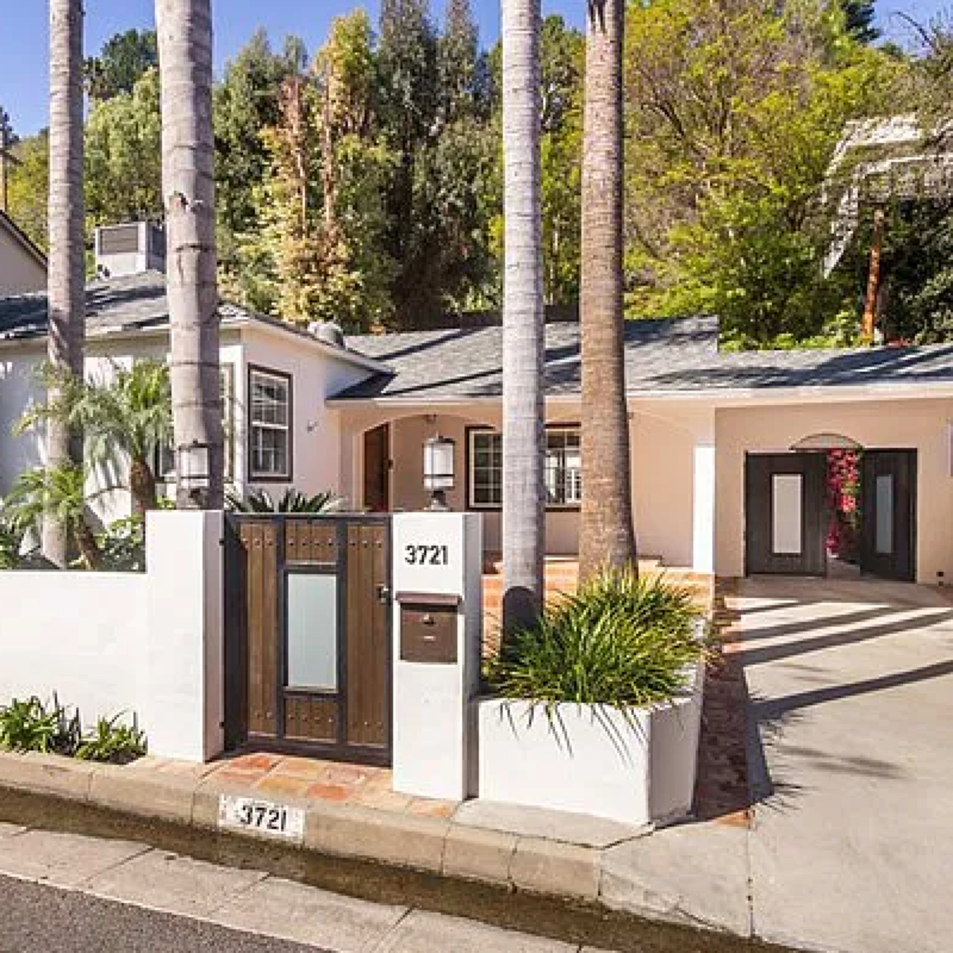 A single-story house with a front yard surrounded by palm trees and a driveway, located on a quiet street.