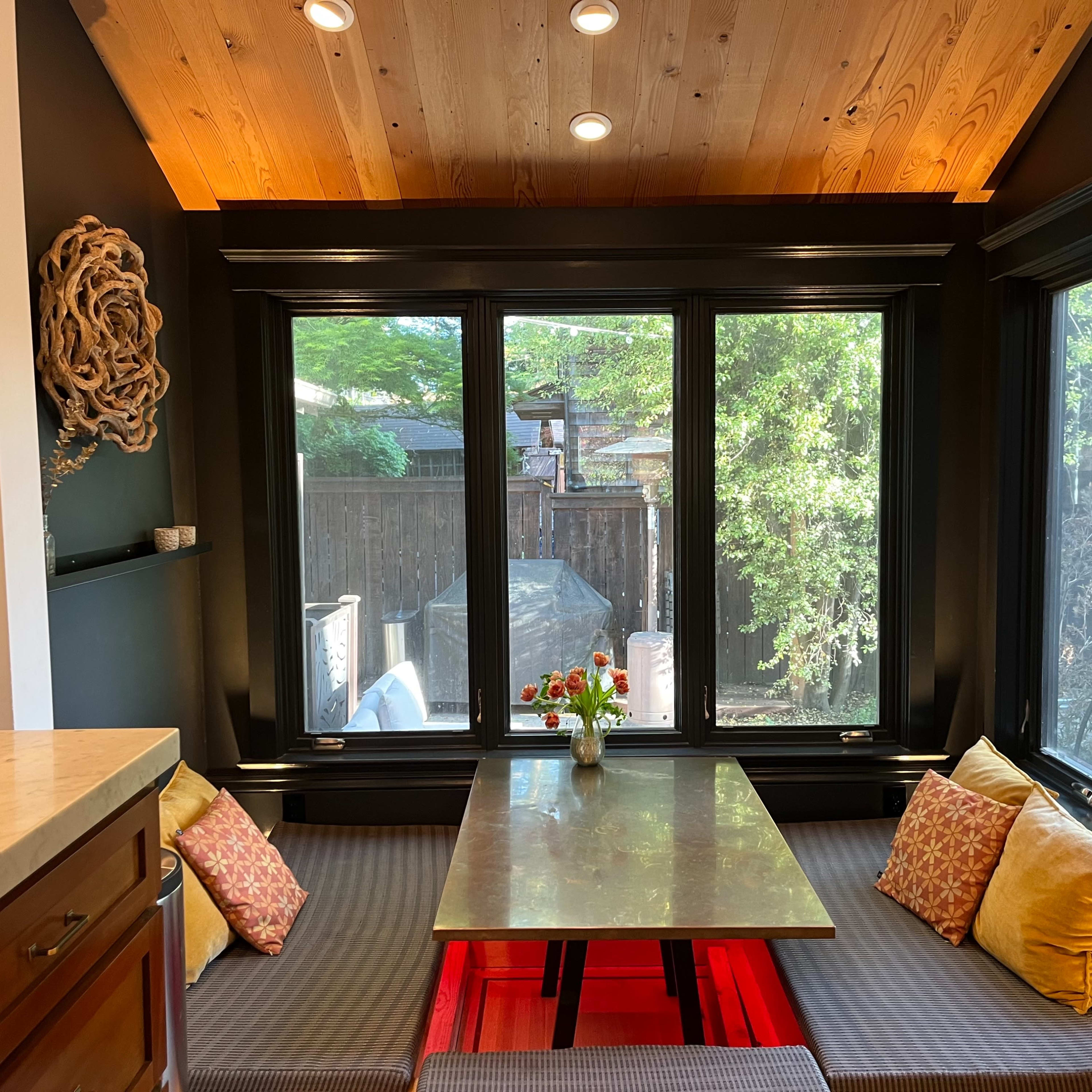 A cozy dining nook with a wooden table surrounded by cushioned bench seating, situated next to large windows that offer a view of greenery outside.