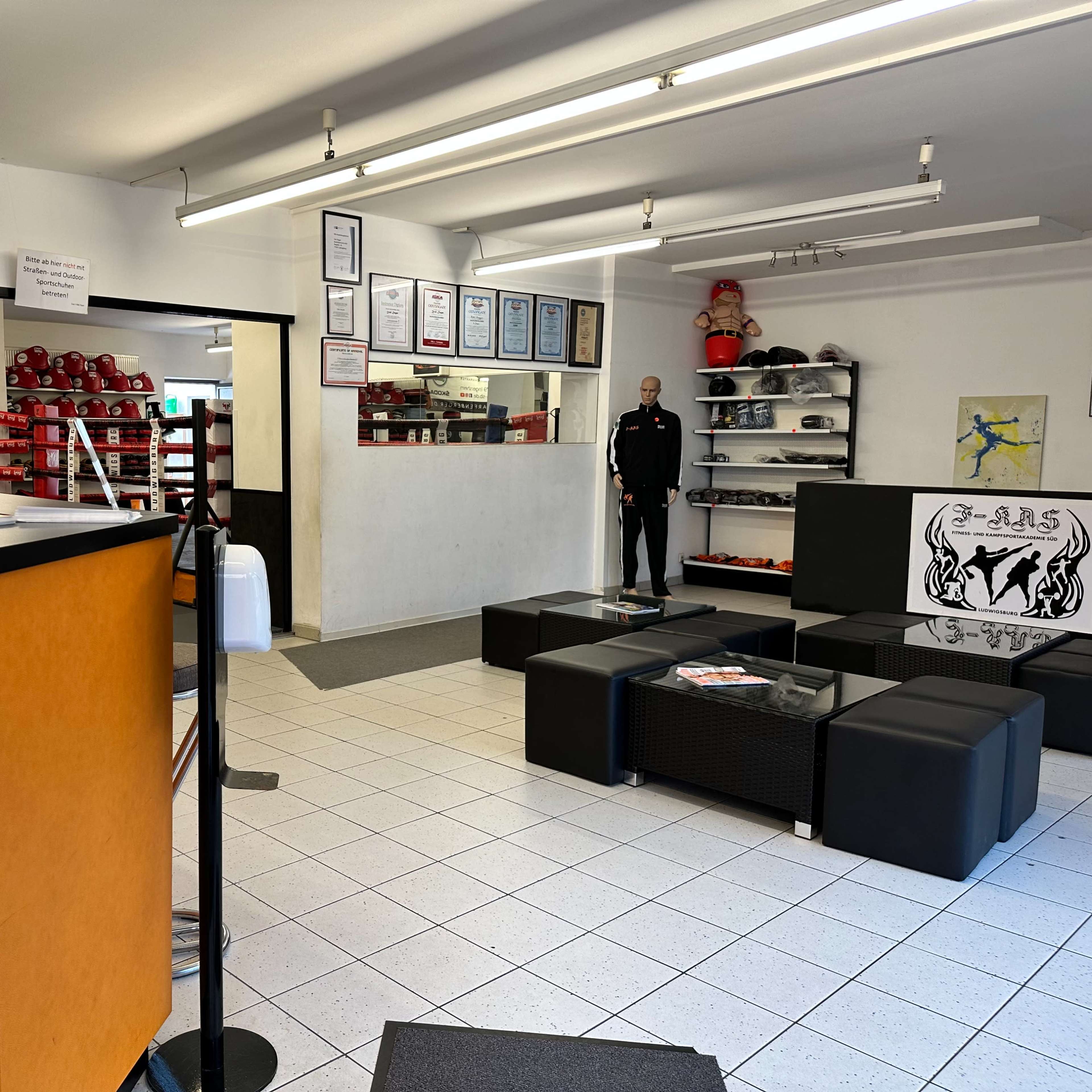 An indoor space with a reception area, featuring an orange counter, black seating, and a wall displaying various certificates and boxing equipment.