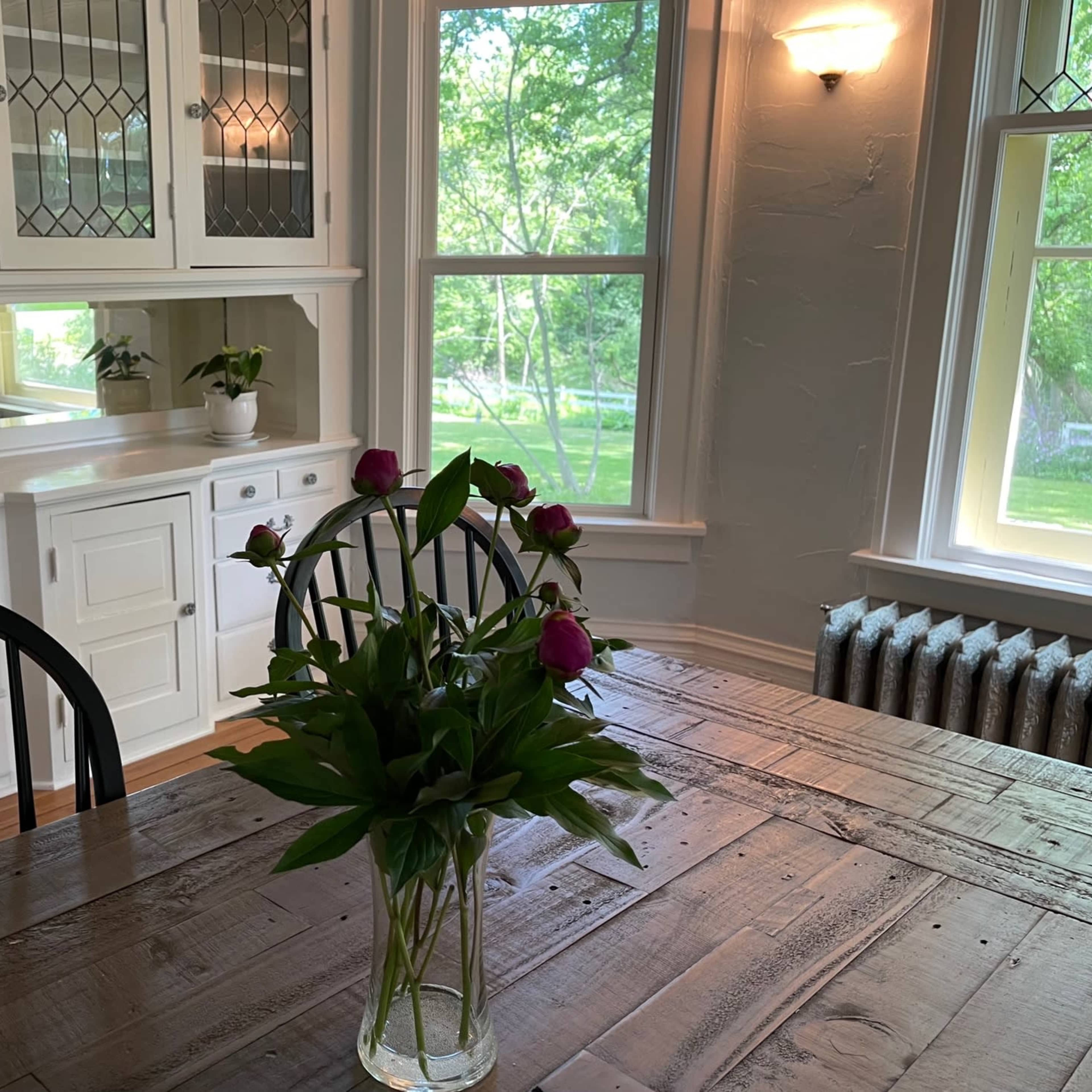 A dining area features a wooden table with a vase of flowers, surrounded by black chairs, and is illuminated by natural light from large windows.