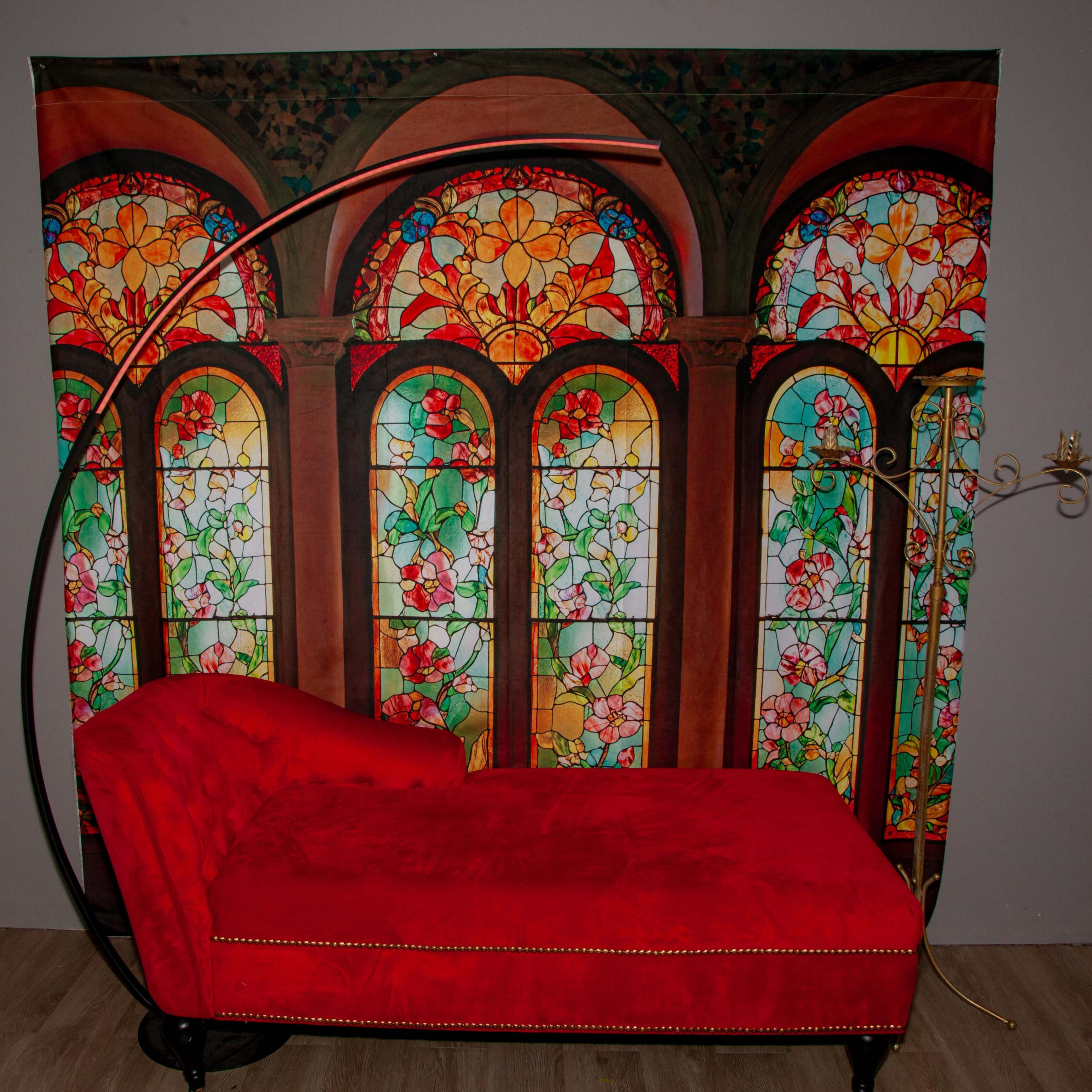 A red chaise lounge is positioned in front of a decorative backdrop featuring stained glass windows with colorful floral designs.