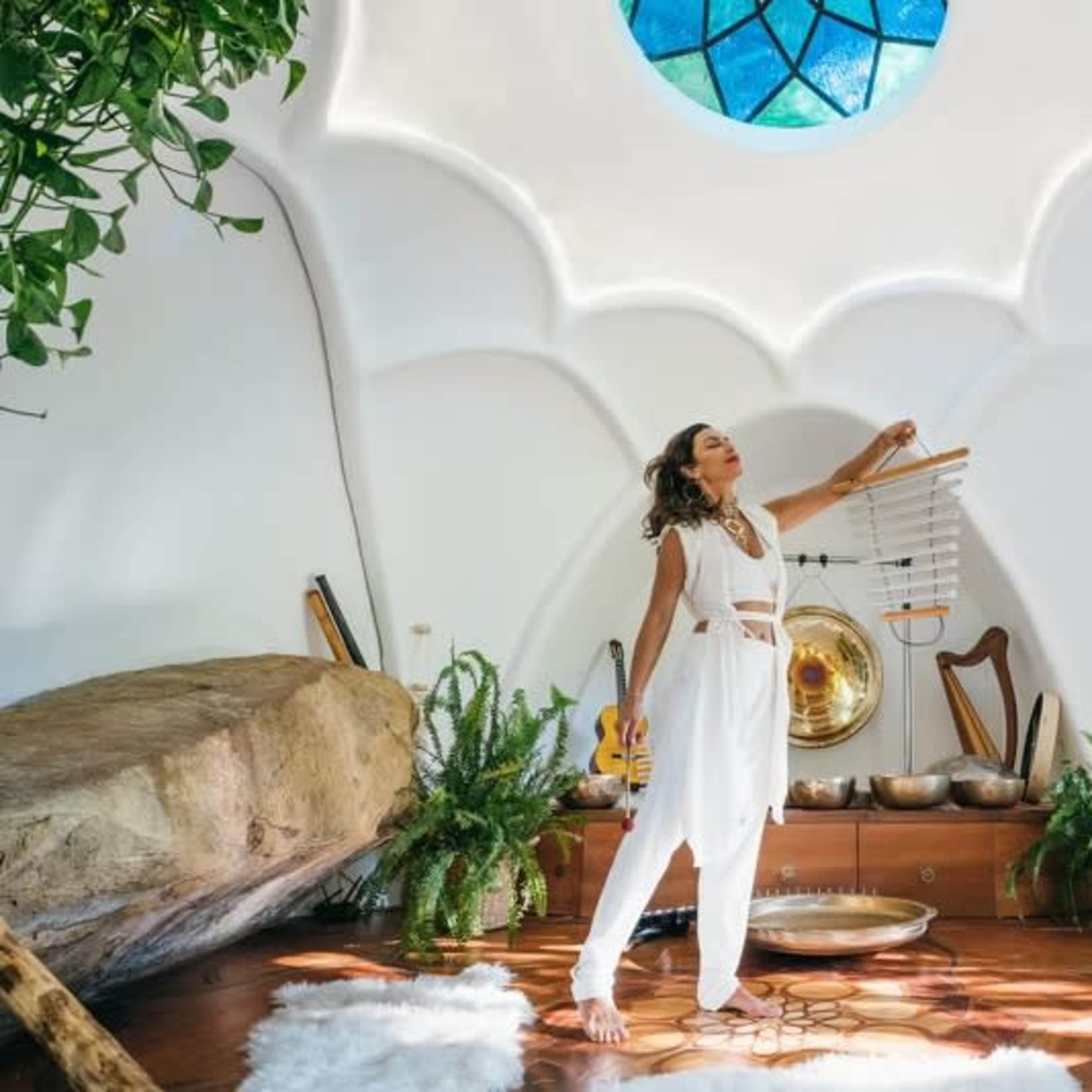 A woman in white clothing stands in a brightly lit, circular room with natural elements and musical instruments.