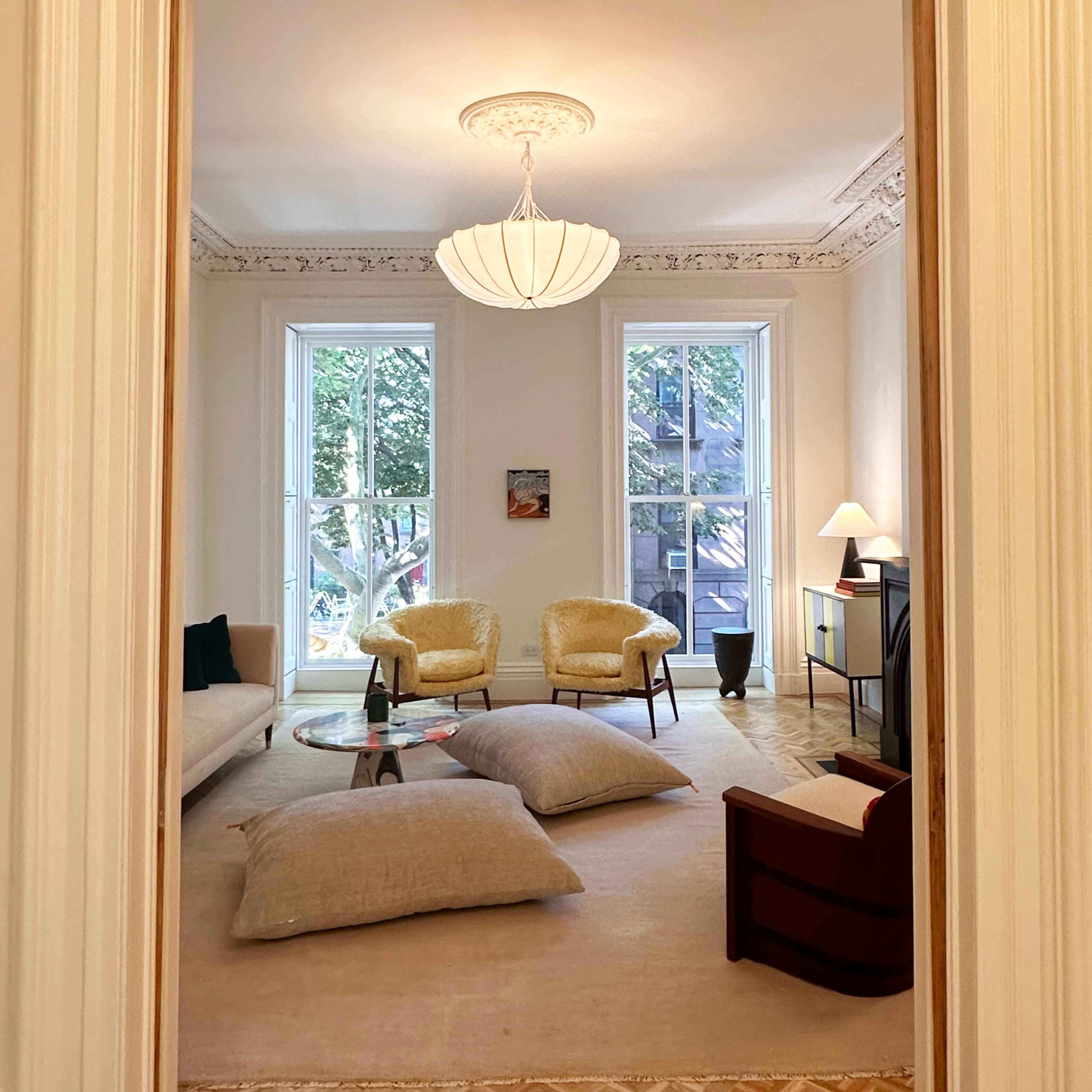 A living room visible through a doorway, featuring a sofa, two yellow chairs, large windows, and a coffee table on a carpeted floor with oversized cushions.