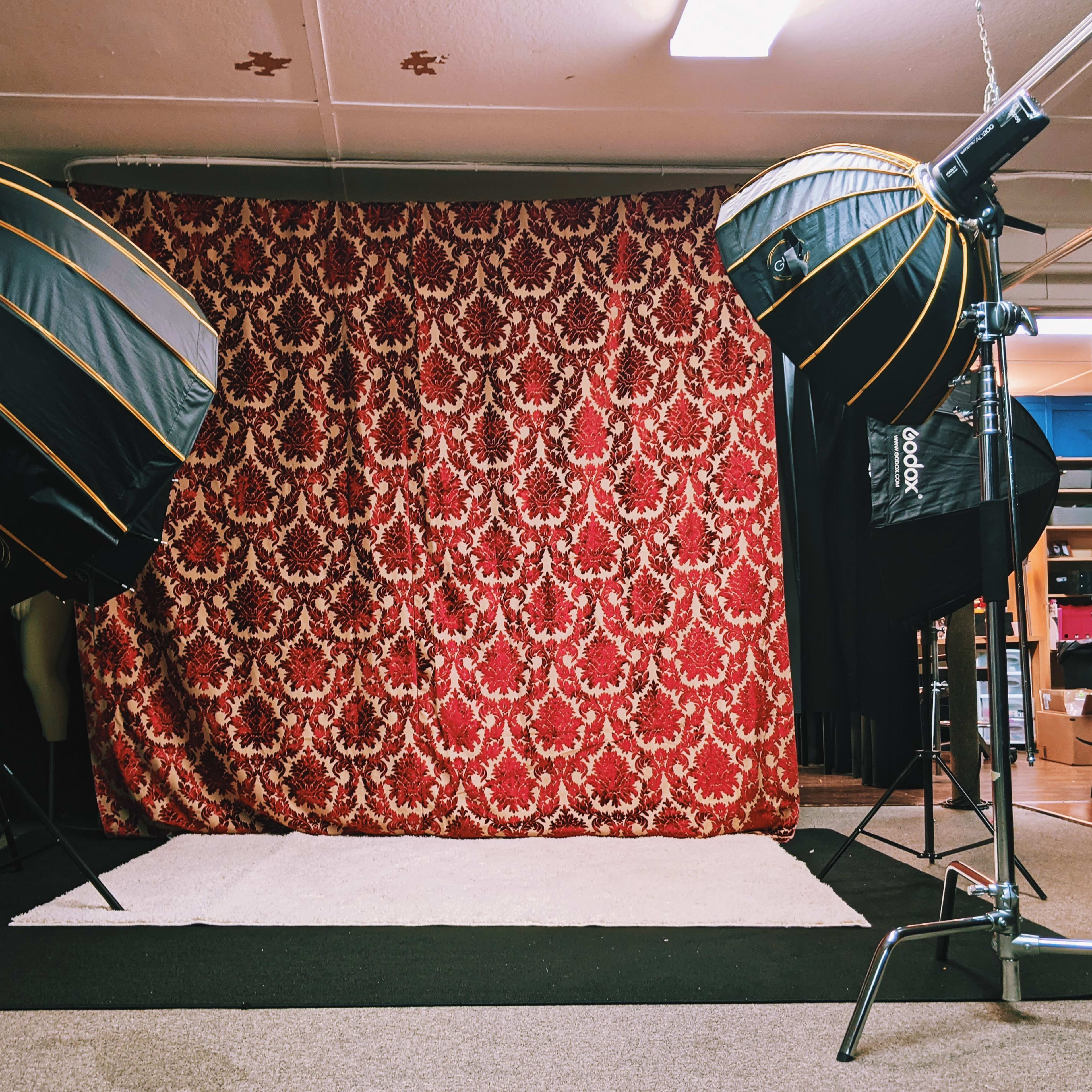 A photography studio setup featuring two large softbox lights positioned in front of a decorative red patterned backdrop and a white rug on the floor.