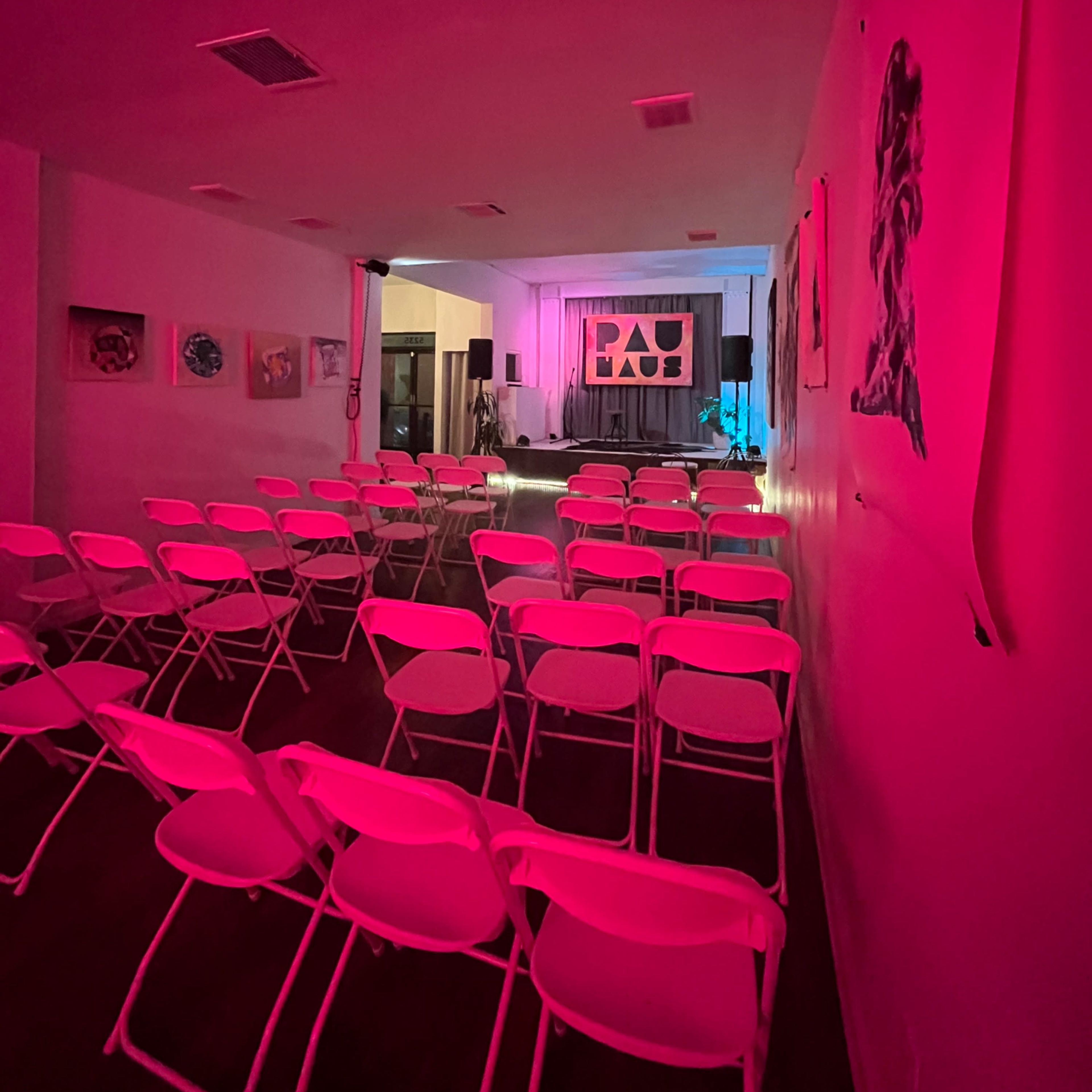A brightly lit event space features neatly arranged white folding chairs facing a stage with a backdrop and artwork on the walls.