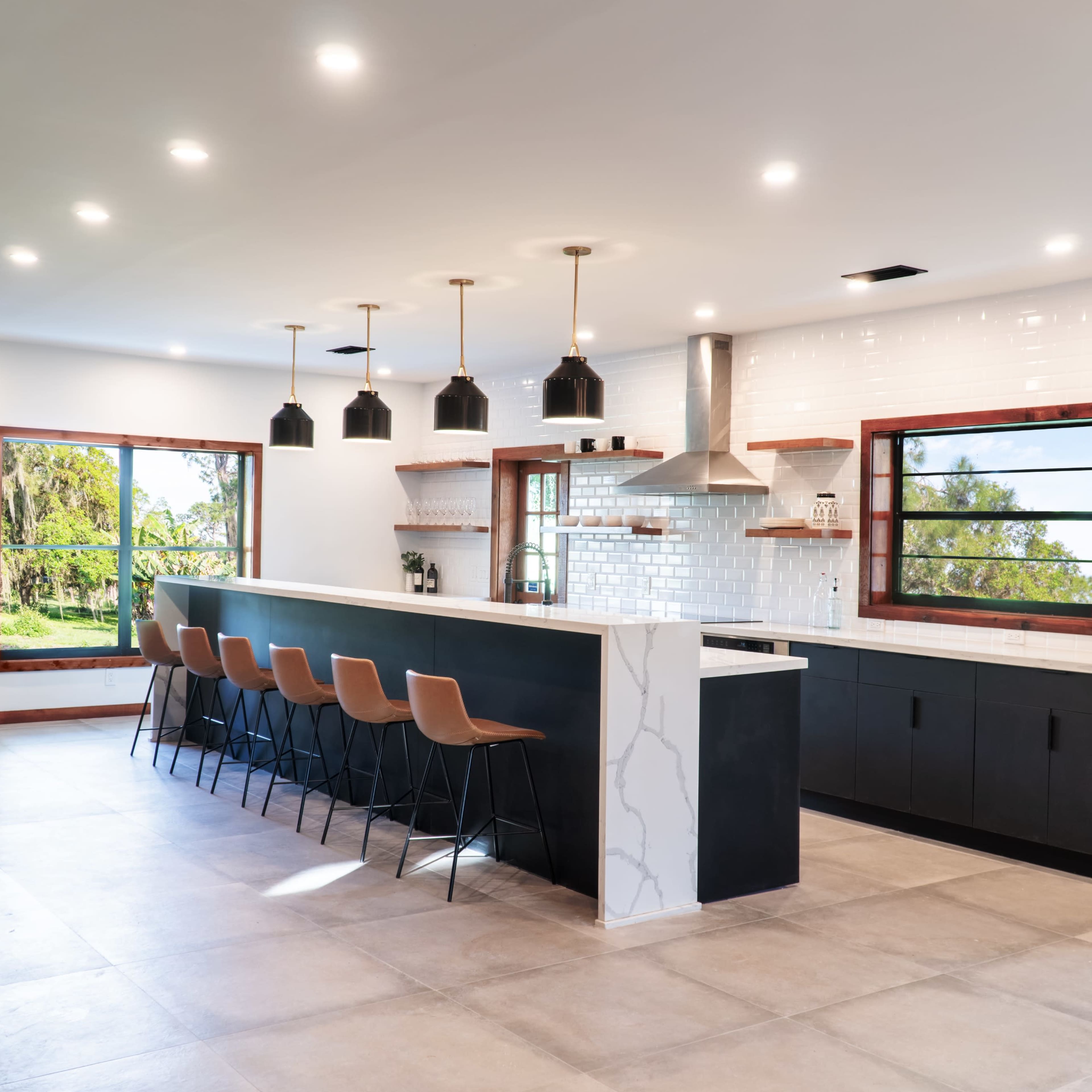 A modern kitchen features a large island with bar seating, pendant lights, and windows that overlook a landscaped area.