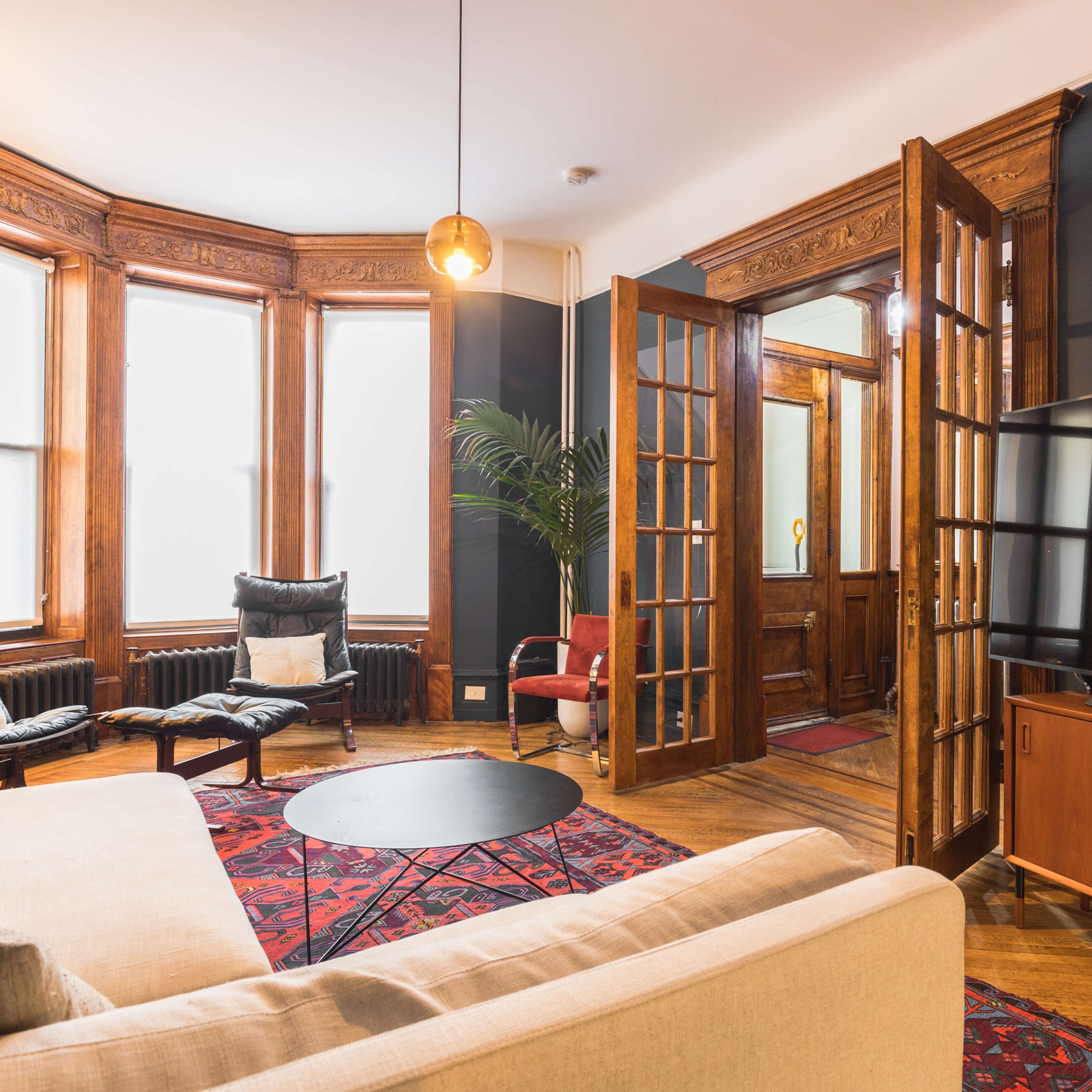 A cozy living room with a beige sofa, black leather chairs, a large television, and decorative wooden details, featuring an entrance with double doors.