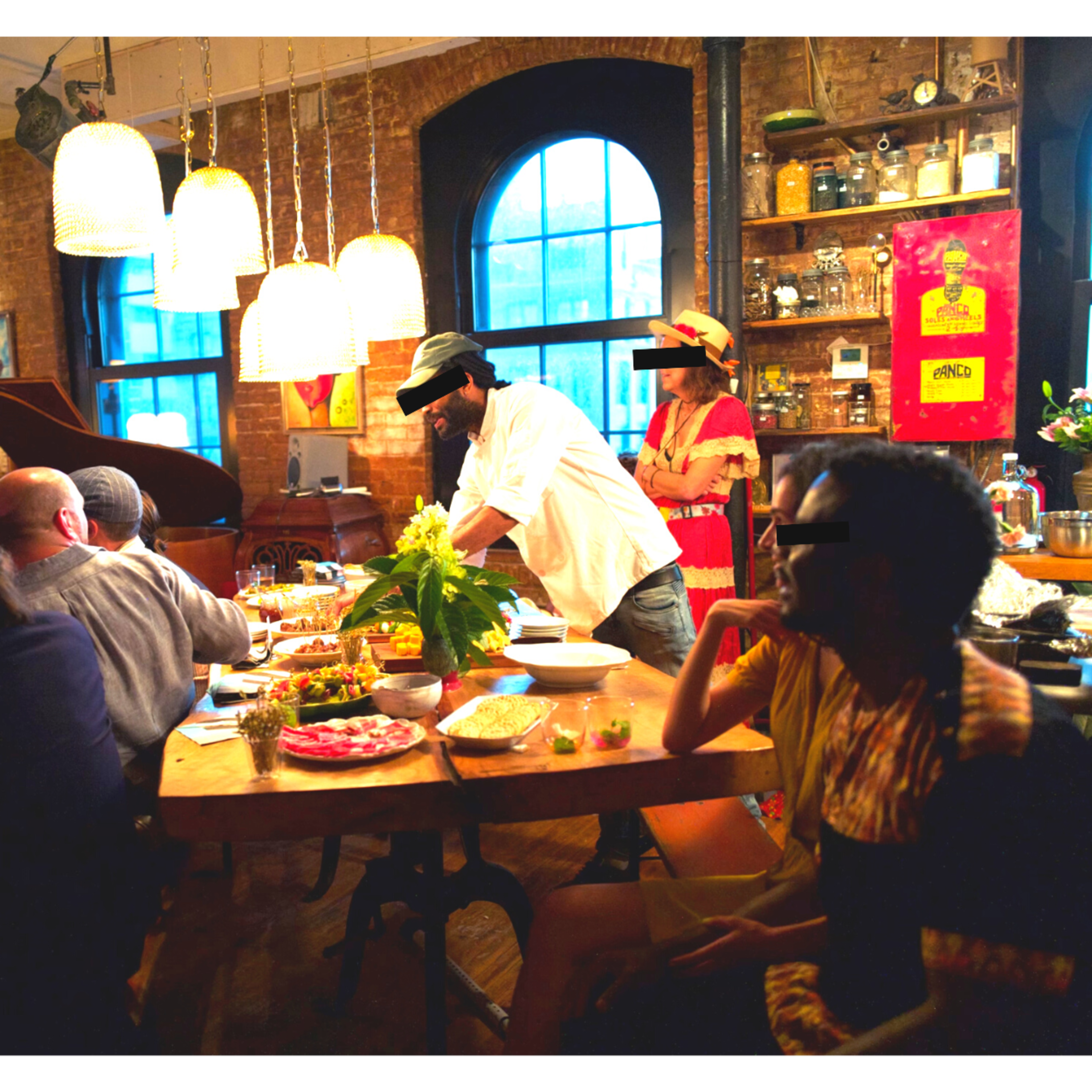 A diverse group of people gathers around a large wooden table filled with food in a warmly lit room featuring brick walls and large windows.