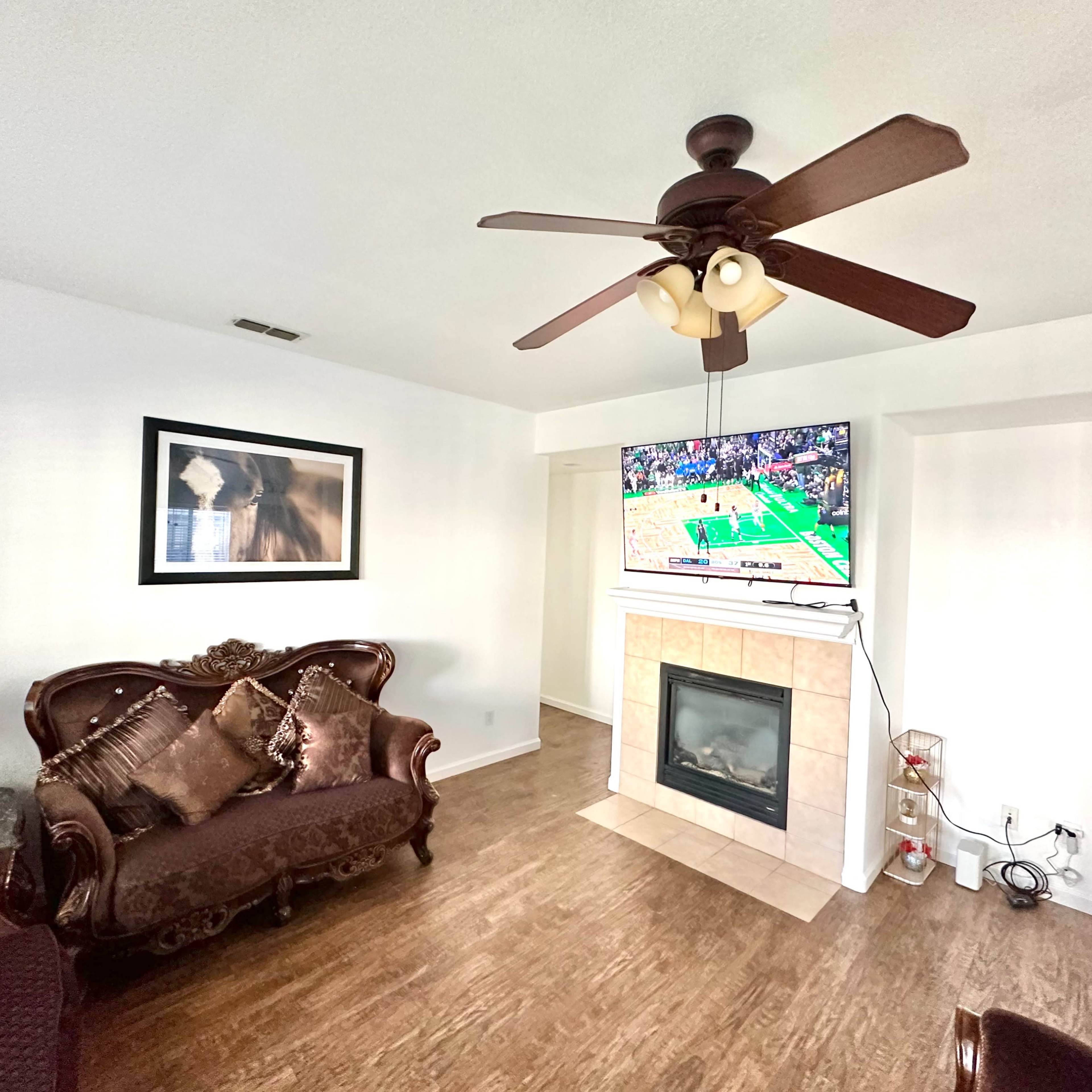 The living room features a brown couch, a mounted television displaying a basketball game, and a fireplace with decorative items nearby.