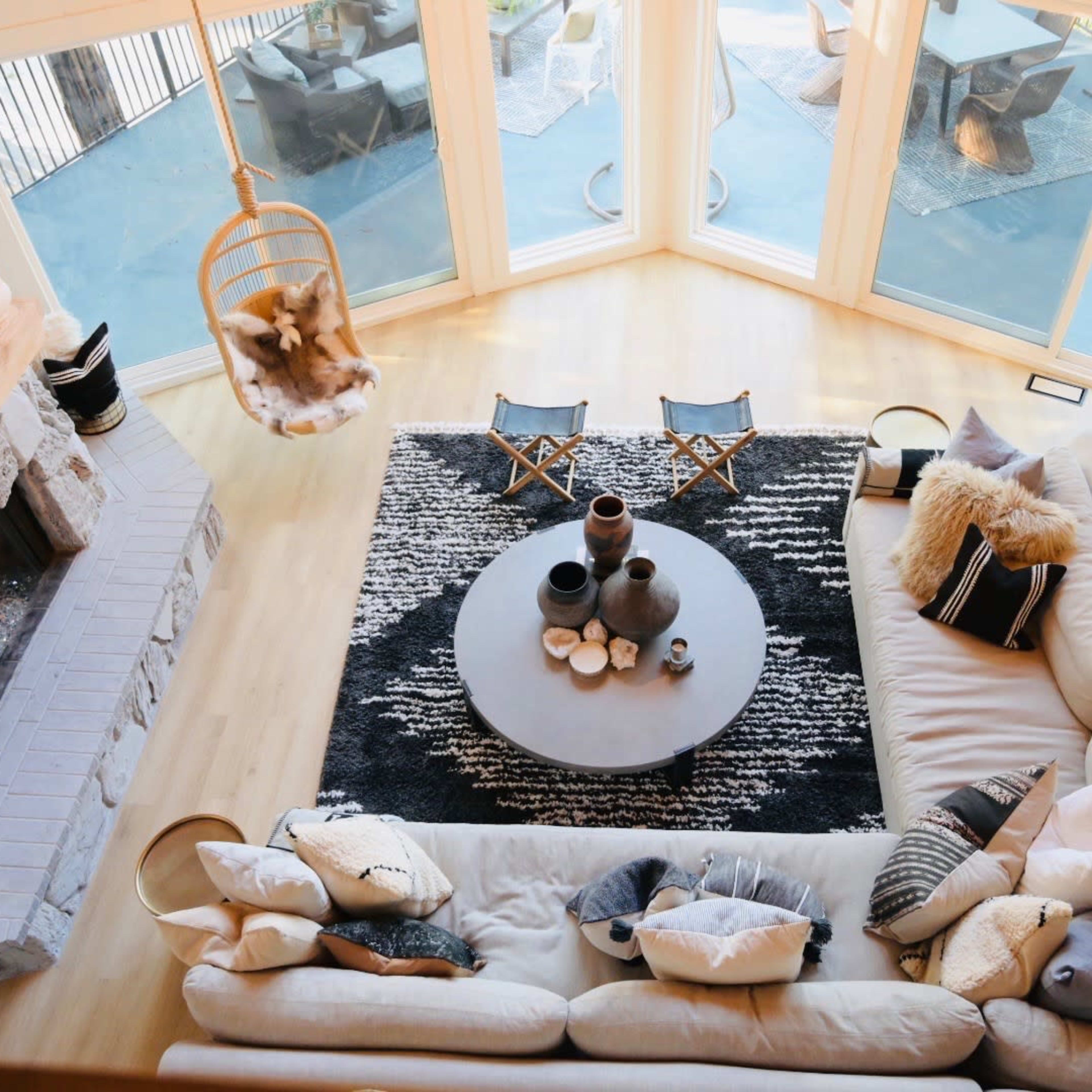A spacious living room features a large sectional sofa, a round coffee table, and a stone fireplace, all arranged around a black and white area rug, with large windows allowing ample natural light.