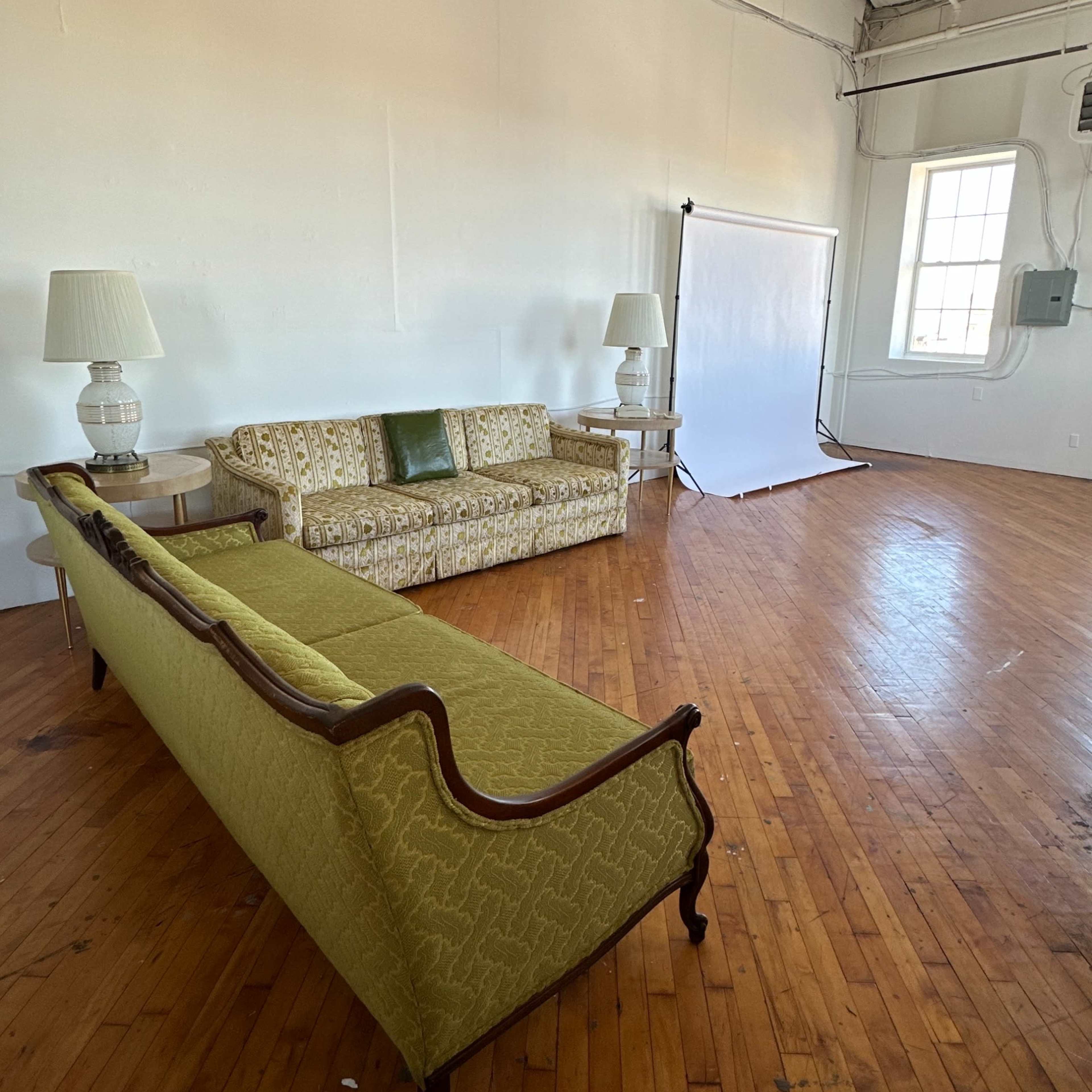 A spacious room with wooden floors featuring a long, ornate green sofa and two matching lamps next to it.