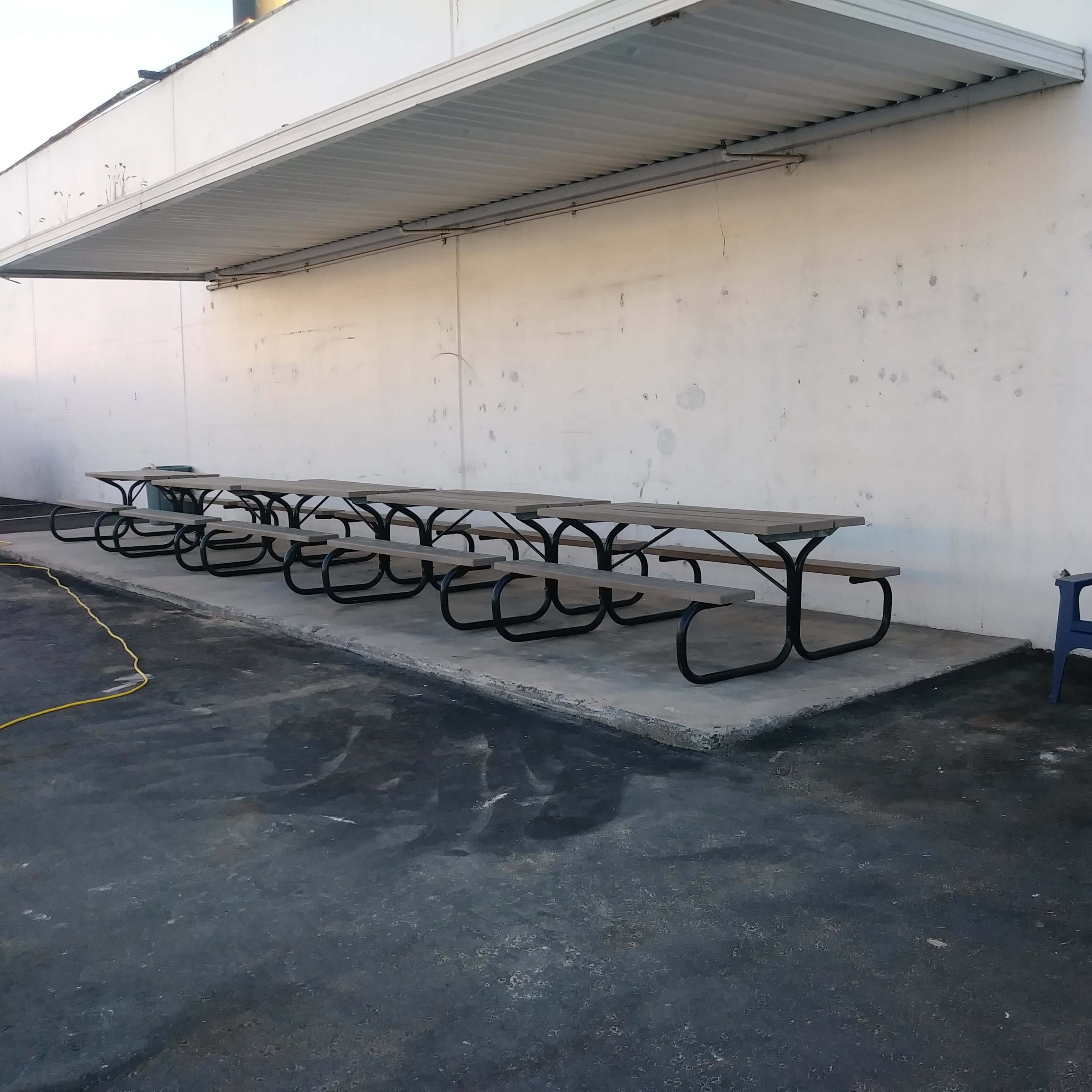 A row of metal picnic tables sits against a white wall beside two blue adirondack chairs.
