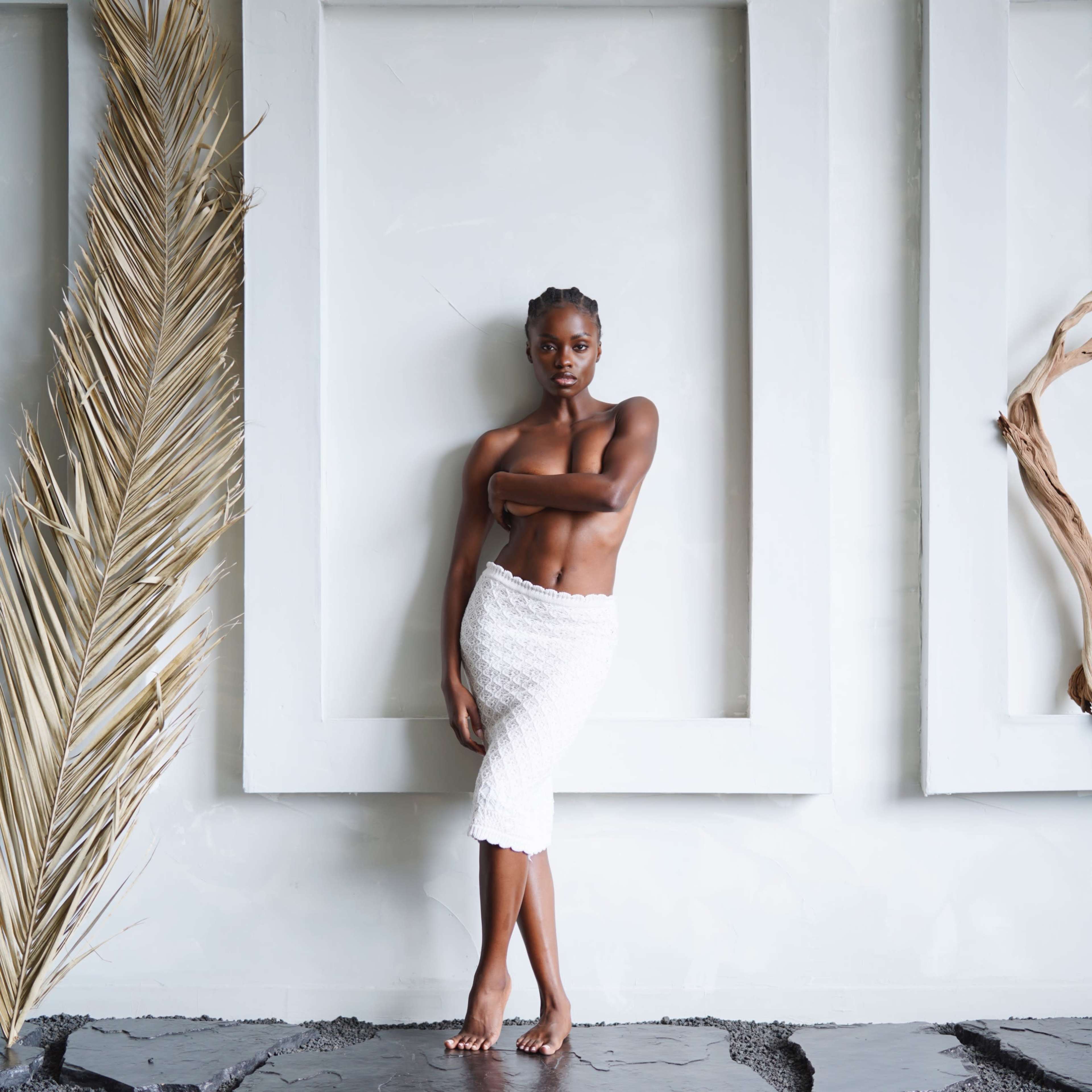 A woman stands barefoot against a textured wall with large, white frames and natural elements nearby.
