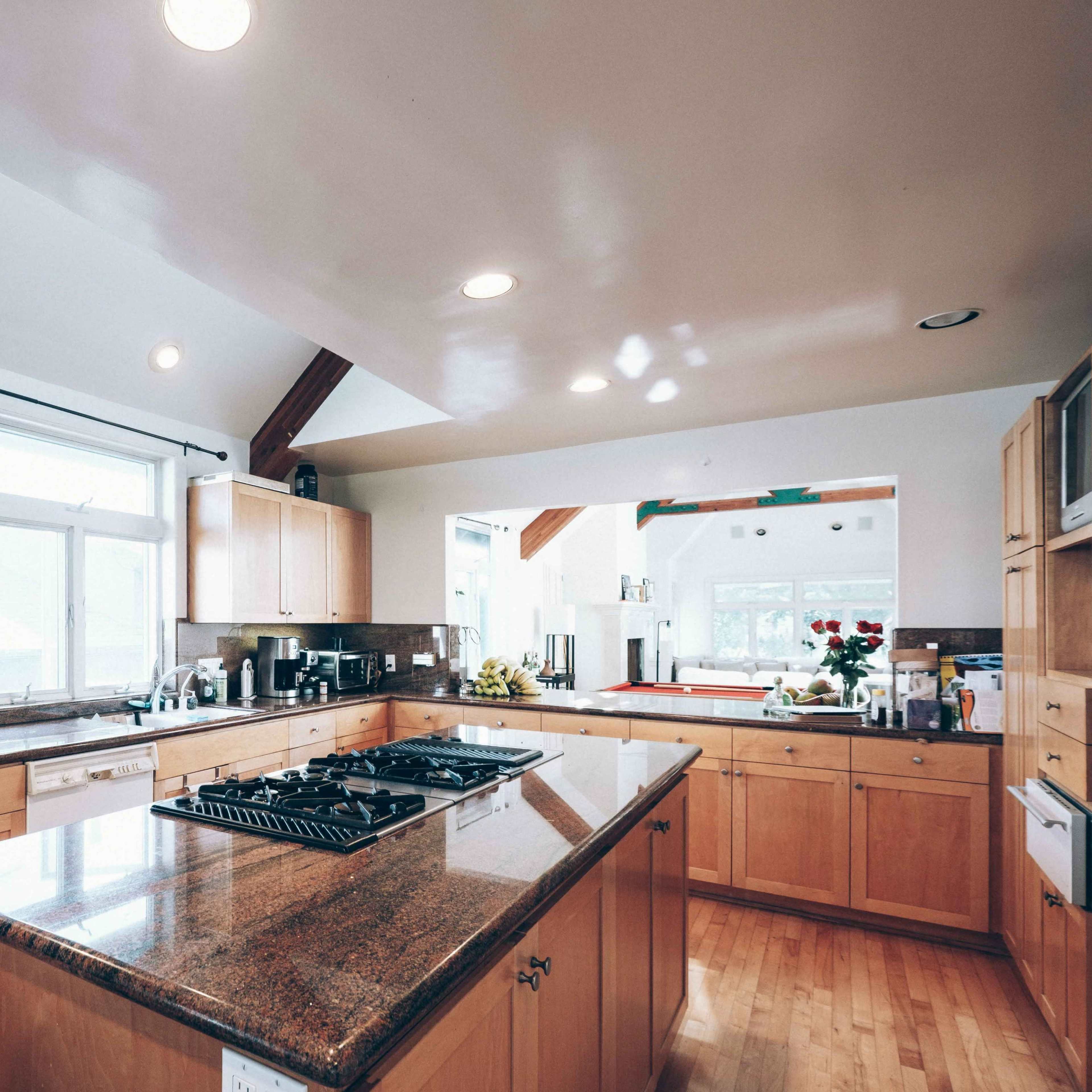 A spacious kitchen with wooden cabinets, a central island with a gas stove, and large windows allowing natural light to enter.
