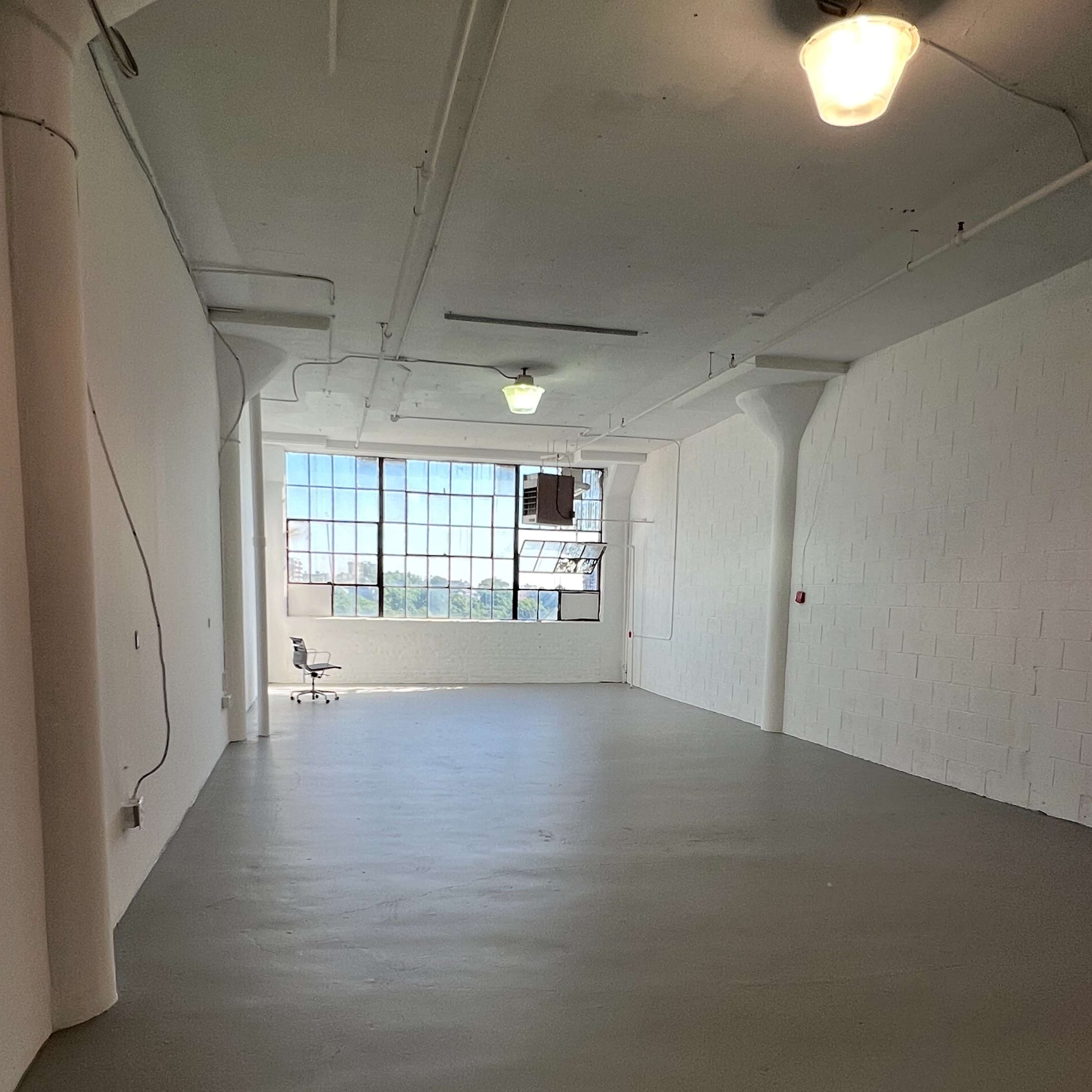 An empty industrial-style room features a large window and a single chair positioned in the corner.