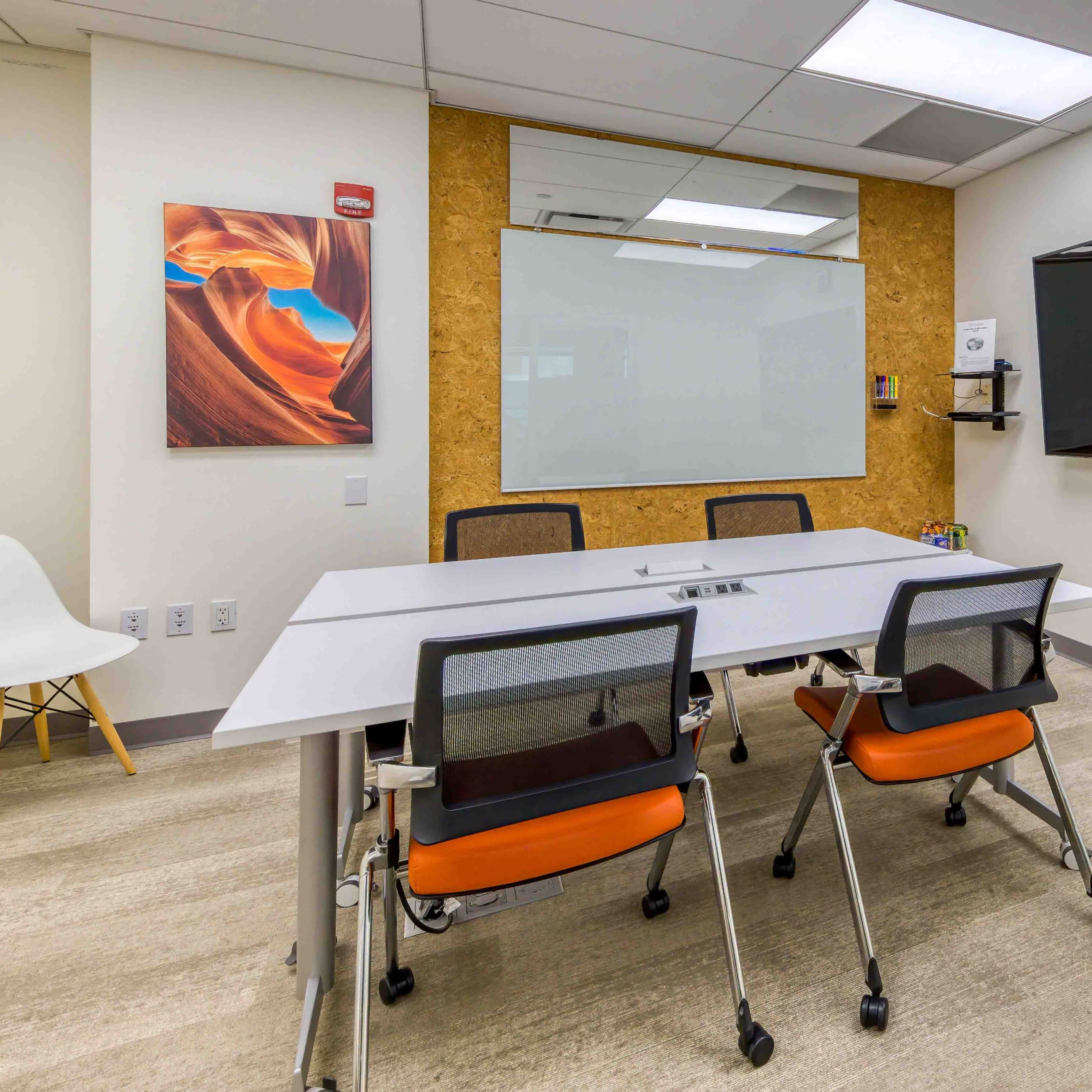 A modern conference room features a white table surrounded by four chairs, a large screen on the wall, a glass whiteboard, and artwork on display.