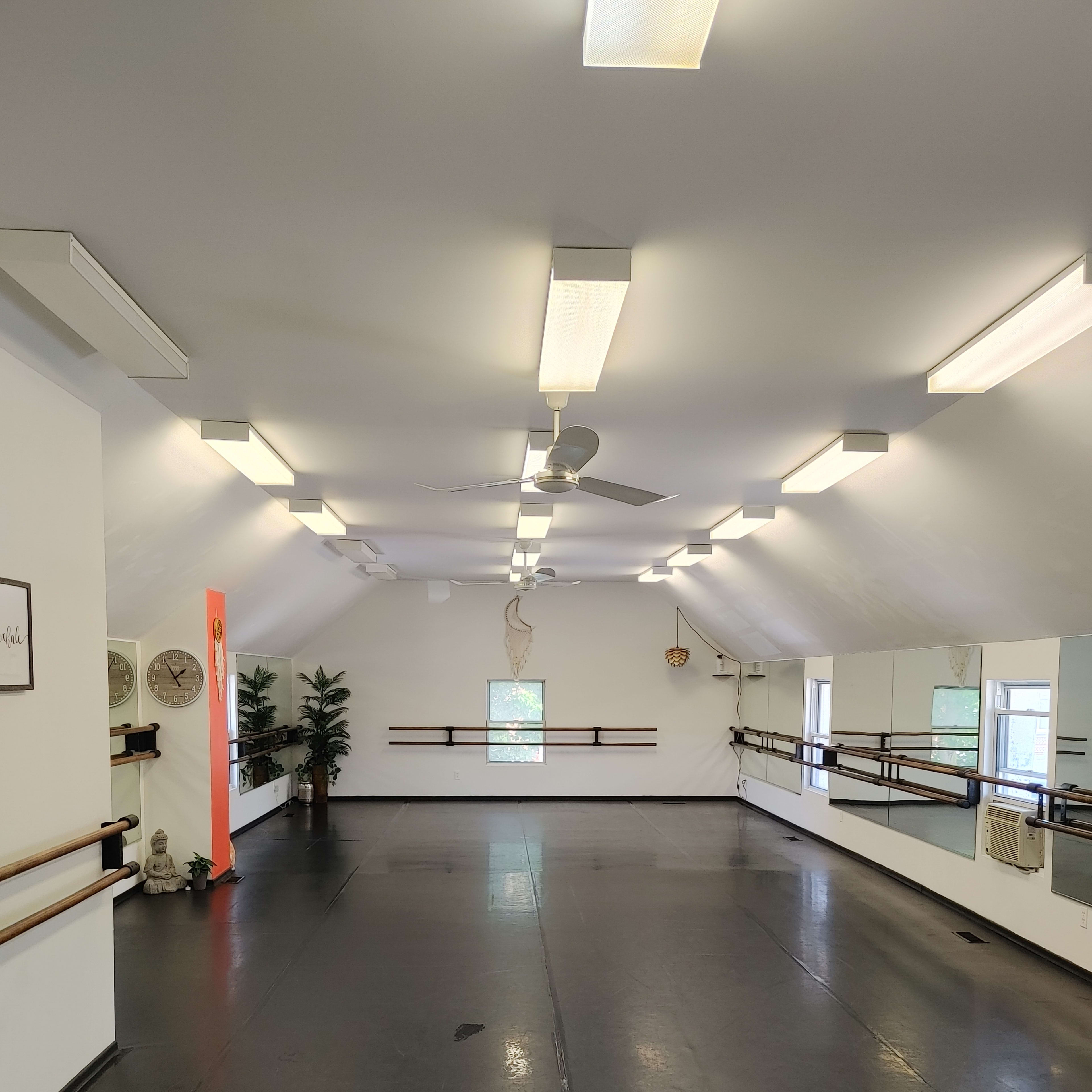 A spacious dance studio with mirrored walls, wooden barre along one side, and bright overhead lights.