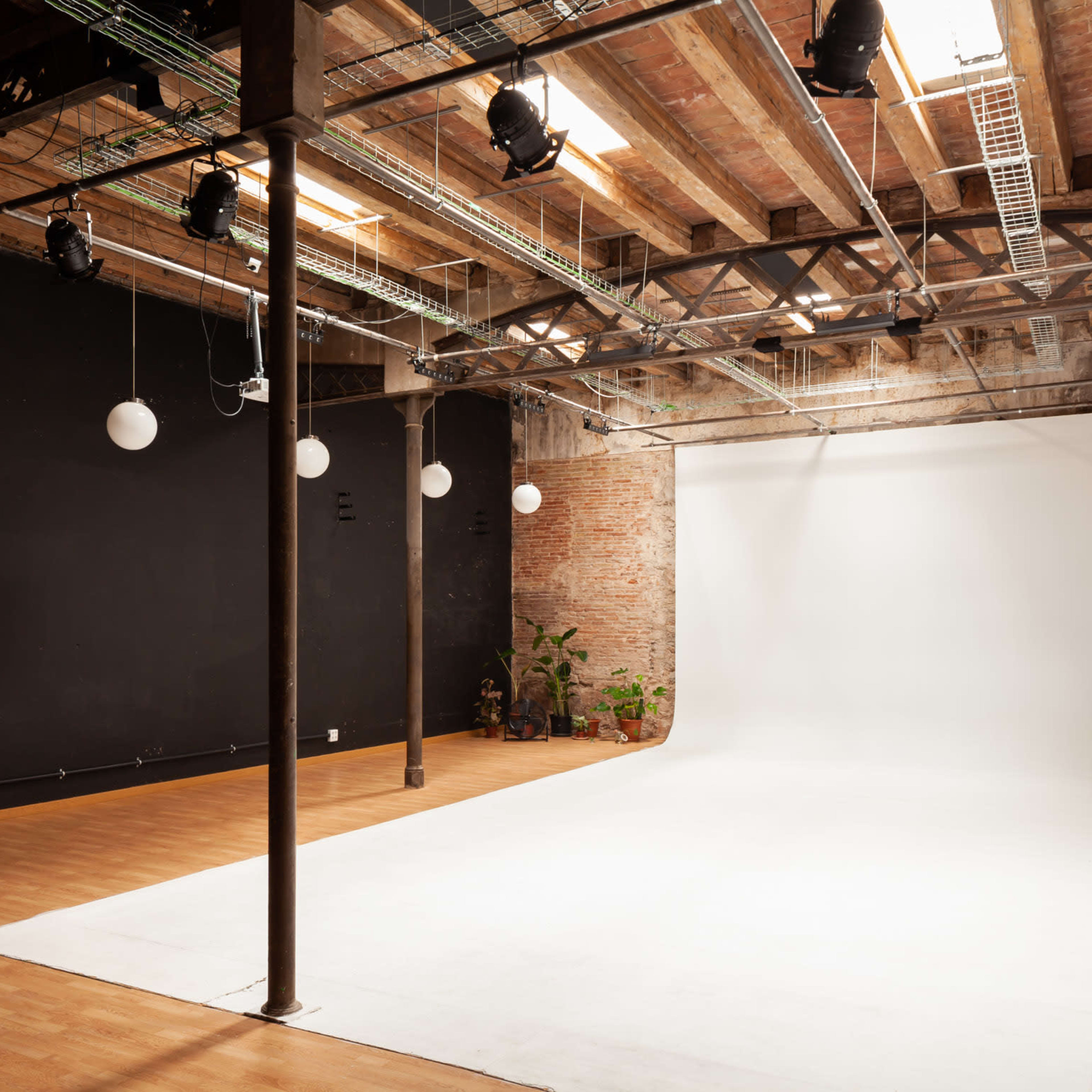 A spacious photography studio with a white backdrop, wooden flooring, exposed brick walls, and overhead lighting.