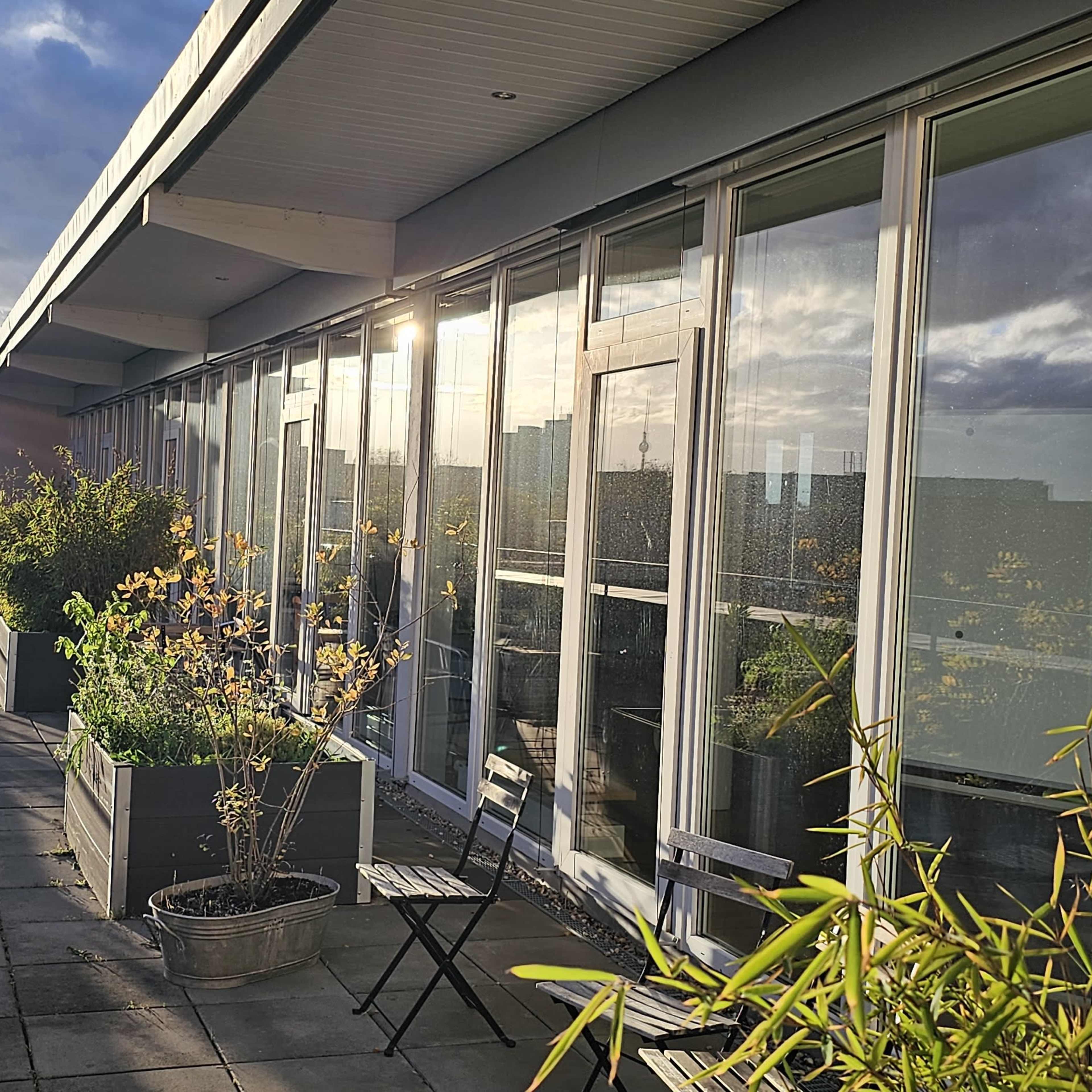 A rooftop terrace with potted plants and chairs, surrounded by glass walls reflecting sunlight.