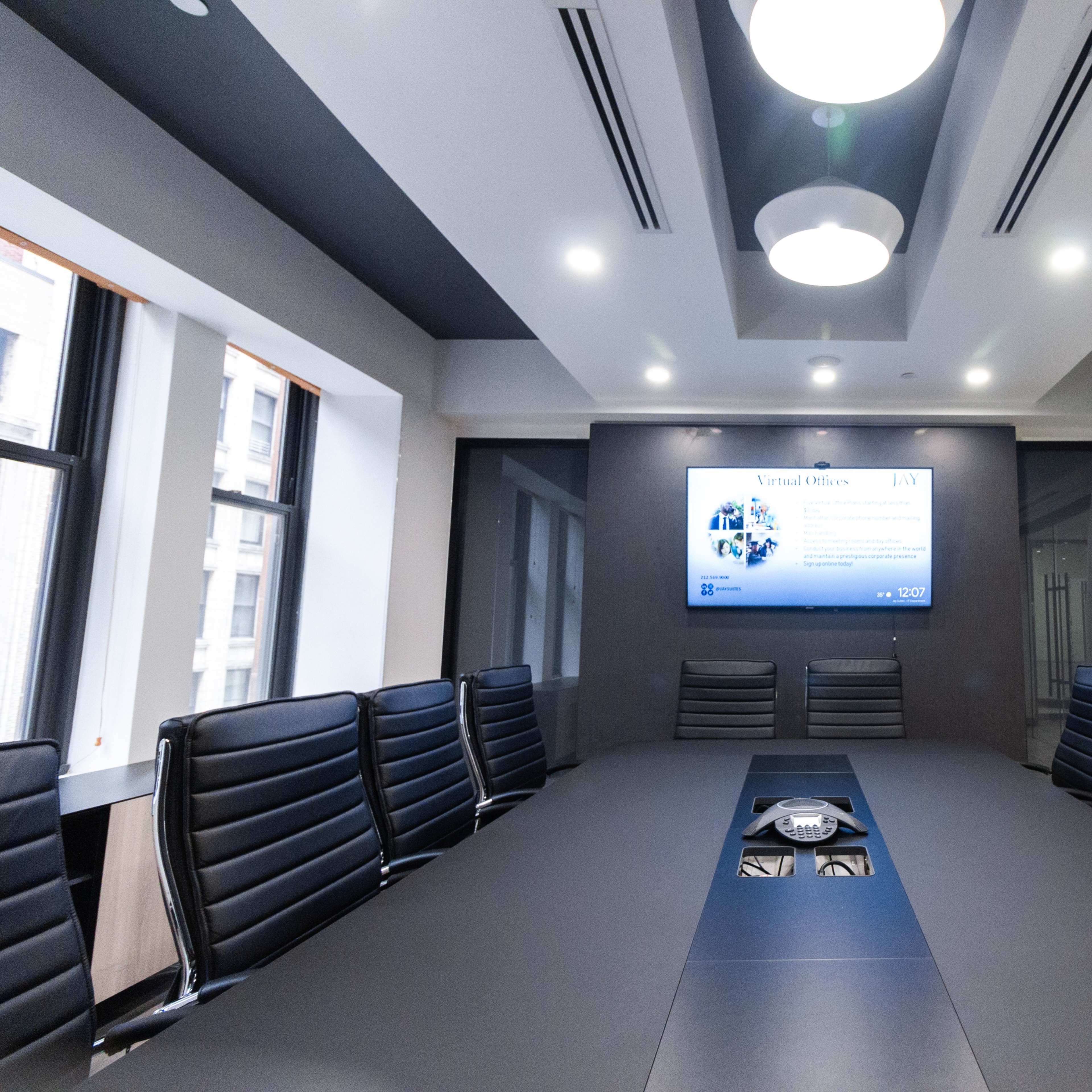 A modern conference room features a long table surrounded by black leather chairs, with a large screen displaying a presentation on the wall.