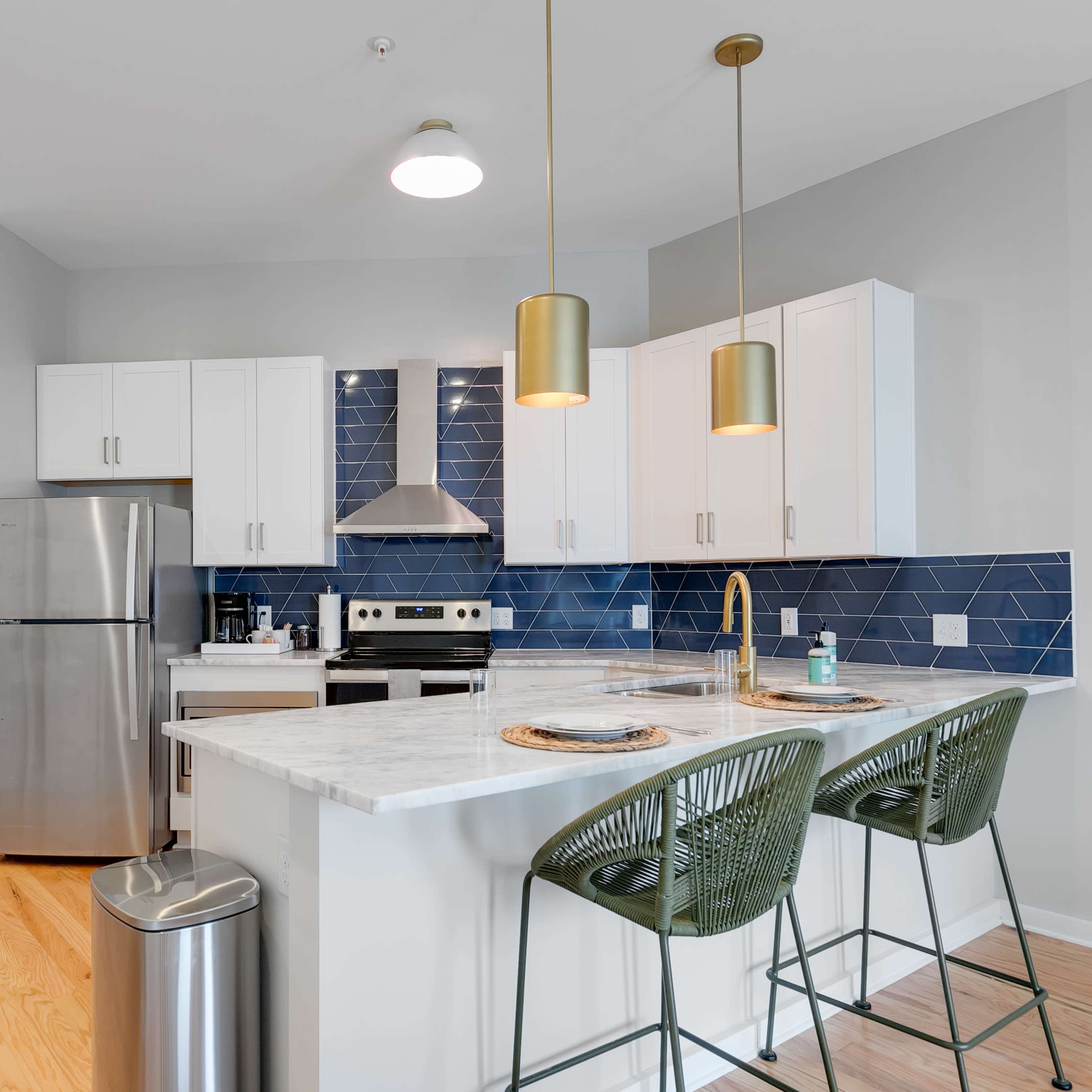 A modern kitchen with stainless steel appliances, white cabinets, a marble countertop, and green bar stools.