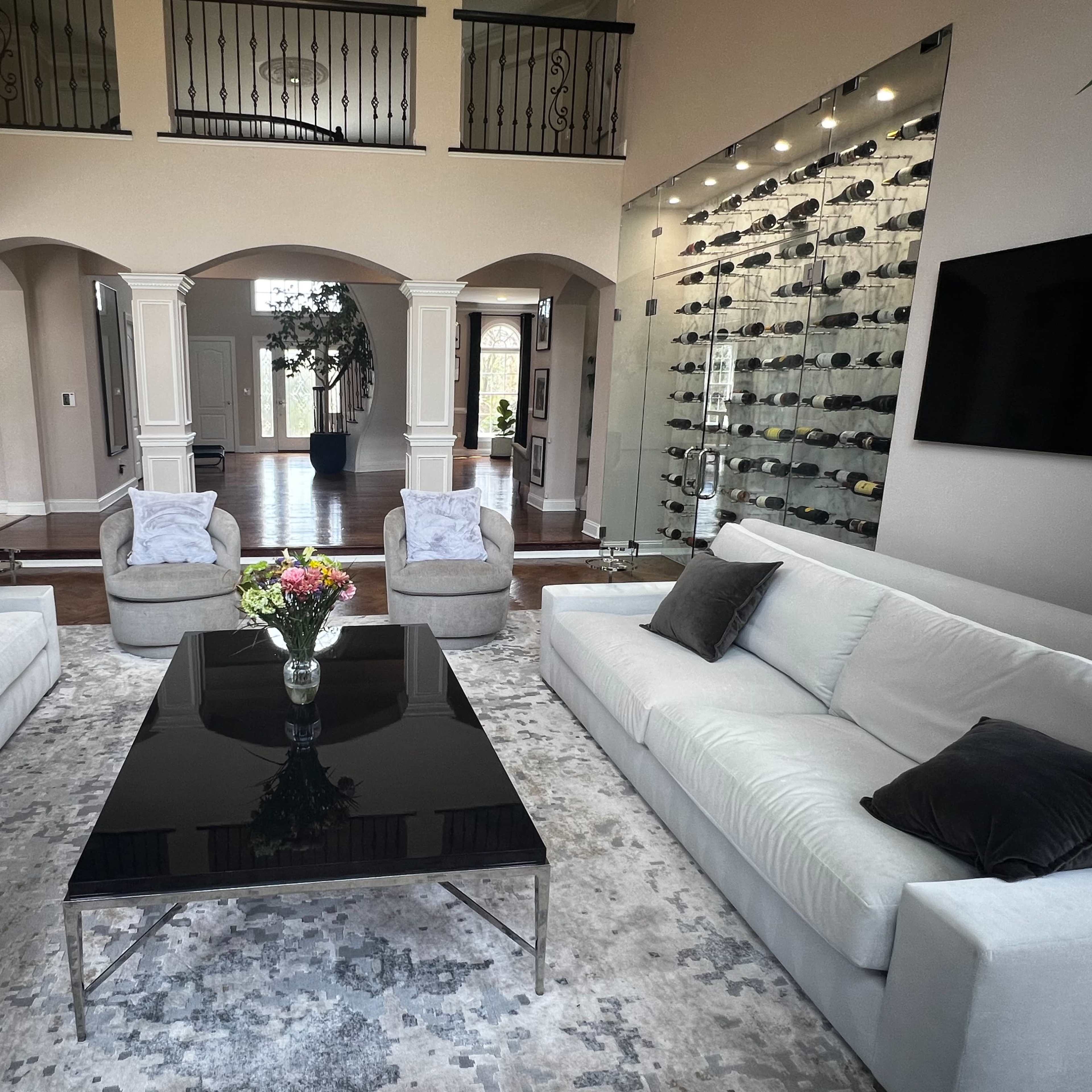 The scene features a spacious living area with a black coffee table at its center, two light gray sofas, two armchairs, and a wall displaying an assortment of wine bottles.