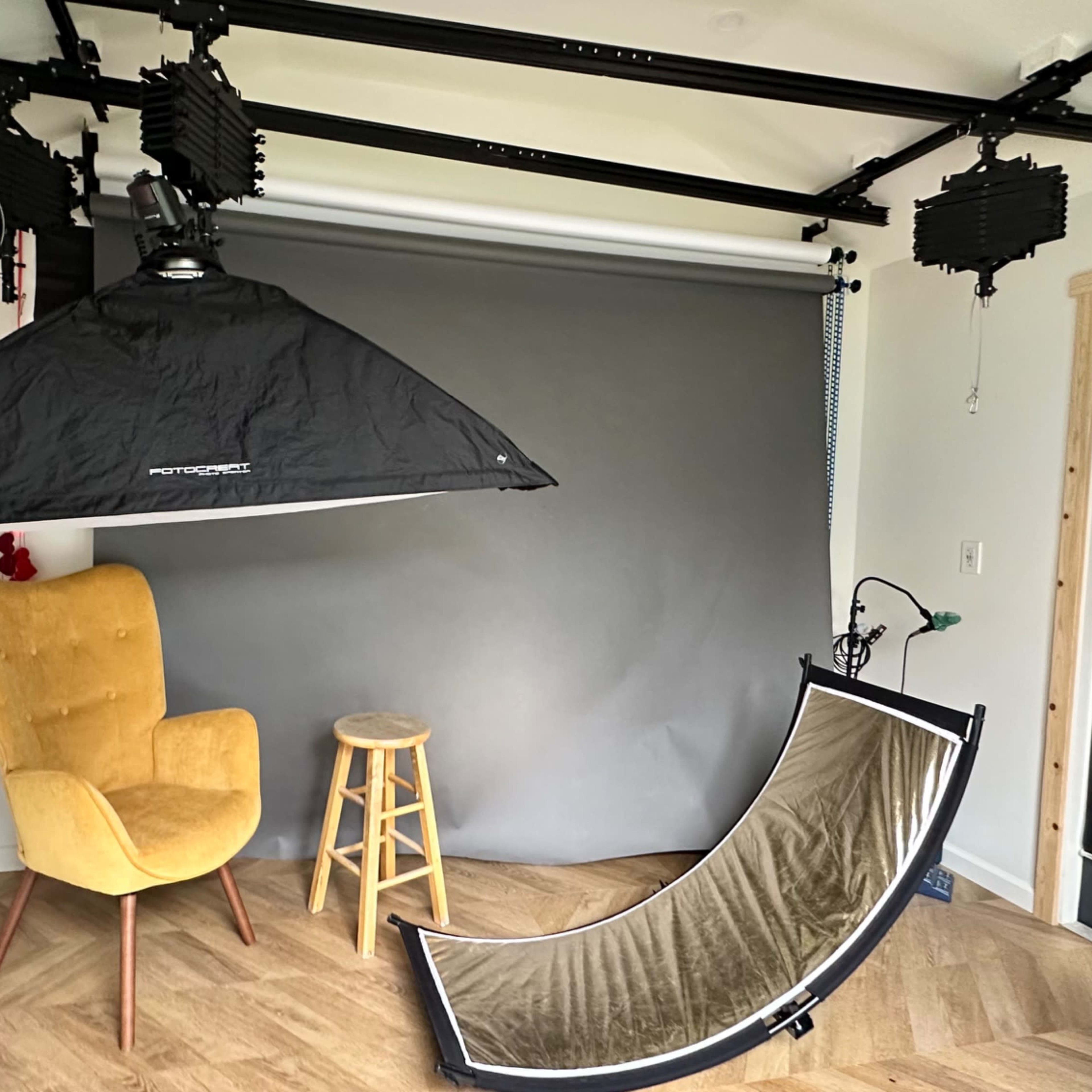 A photography studio setup featuring a gray backdrop, a yellow chair, a wooden stool, and various lighting equipment.