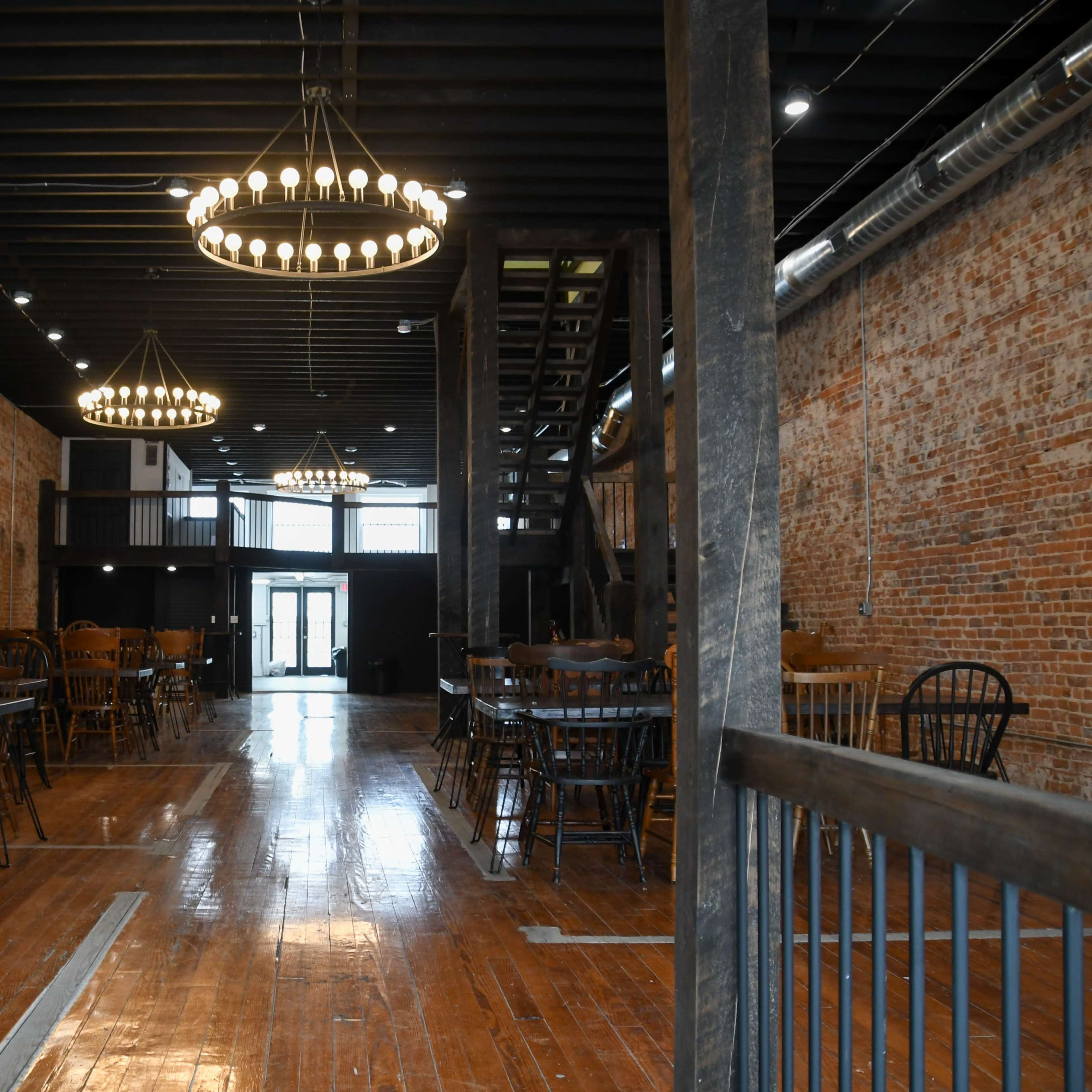 An interior view of a spacious venue featuring wooden flooring, brick walls, and multiple chandeliers hanging from the ceiling.