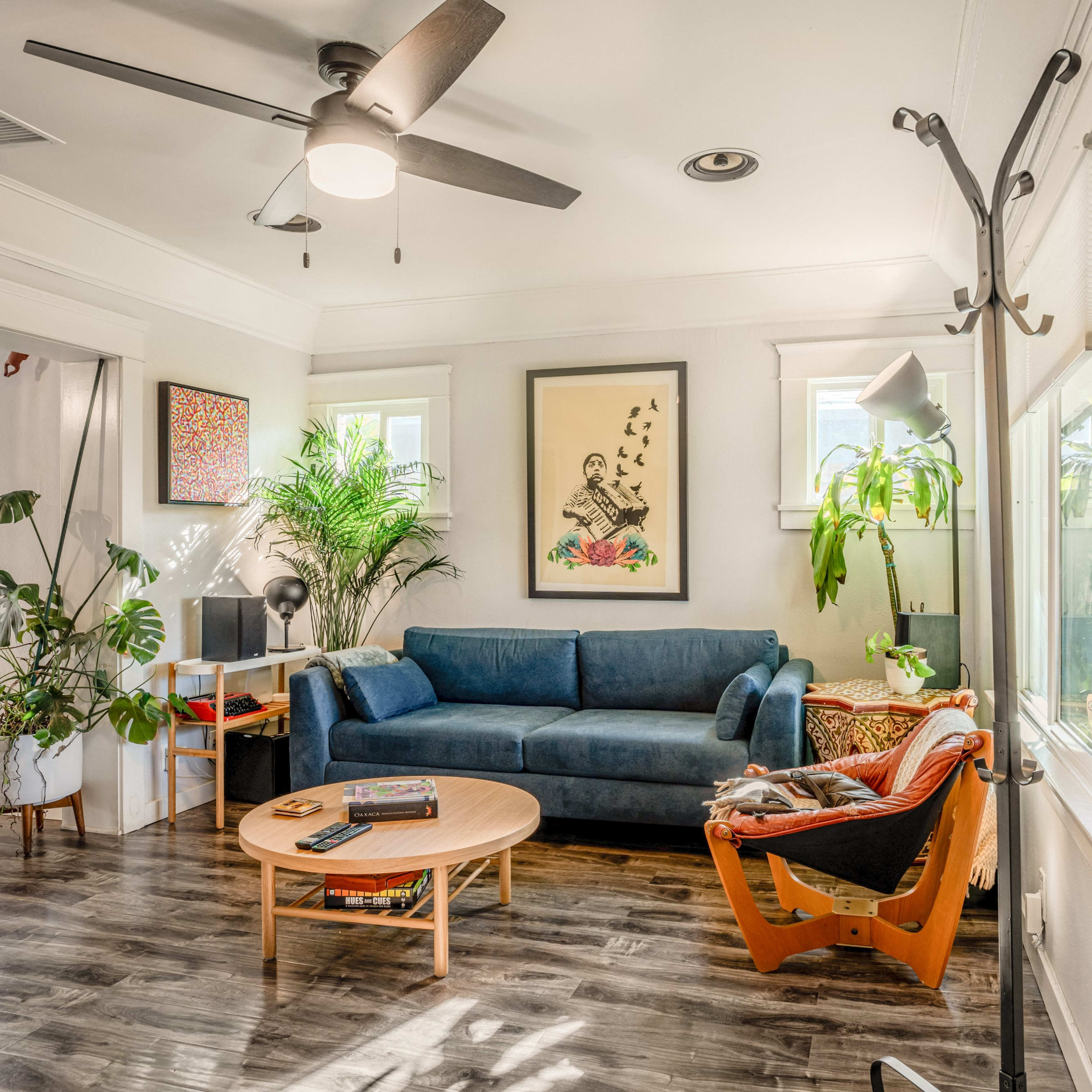 A living room with a blue sofa, a round wooden coffee table, plants, a wall-mounted coat rack, and natural light streaming through the windows.