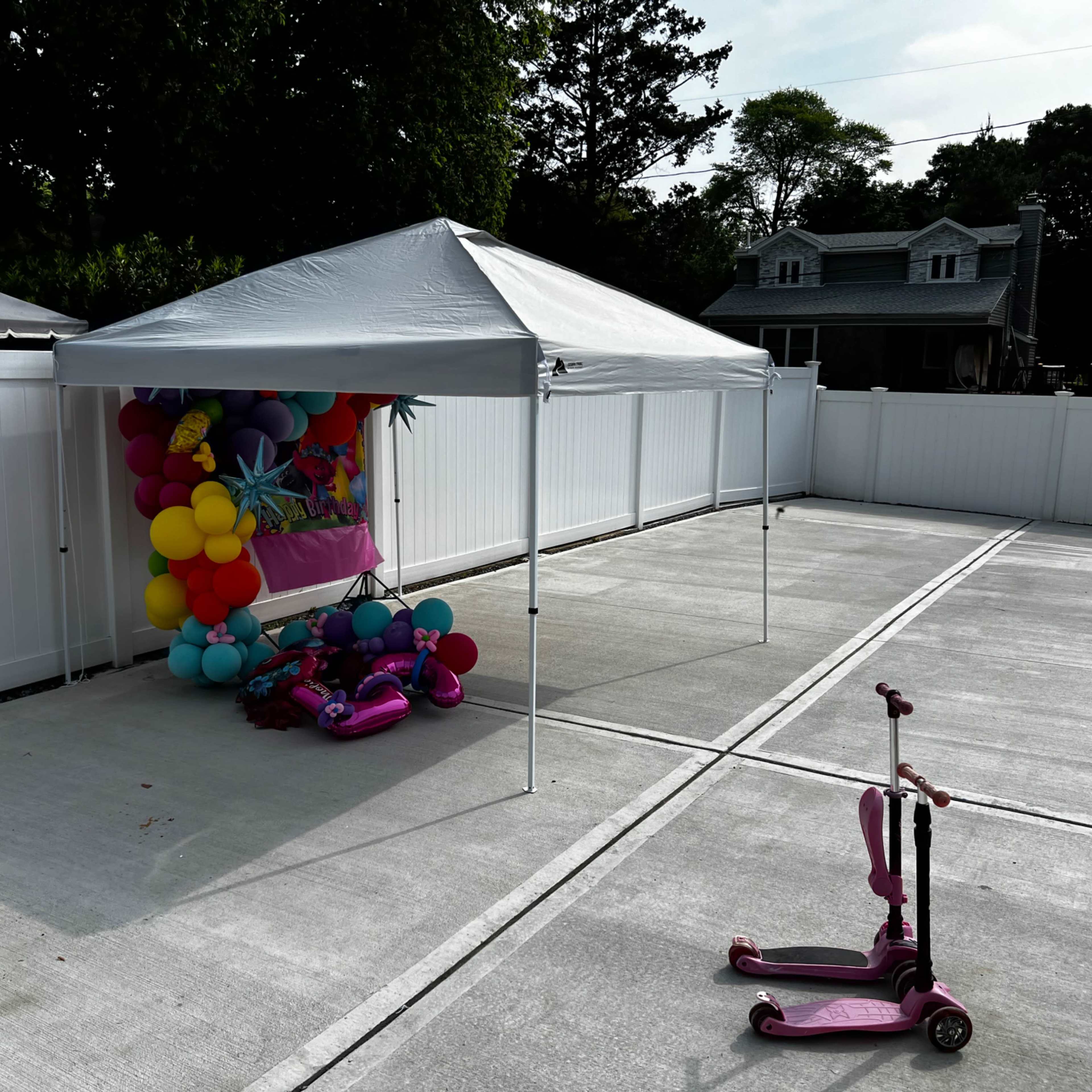 A white canopy tent with colorful balloons is set up in an empty concrete area beside two pink scooters.