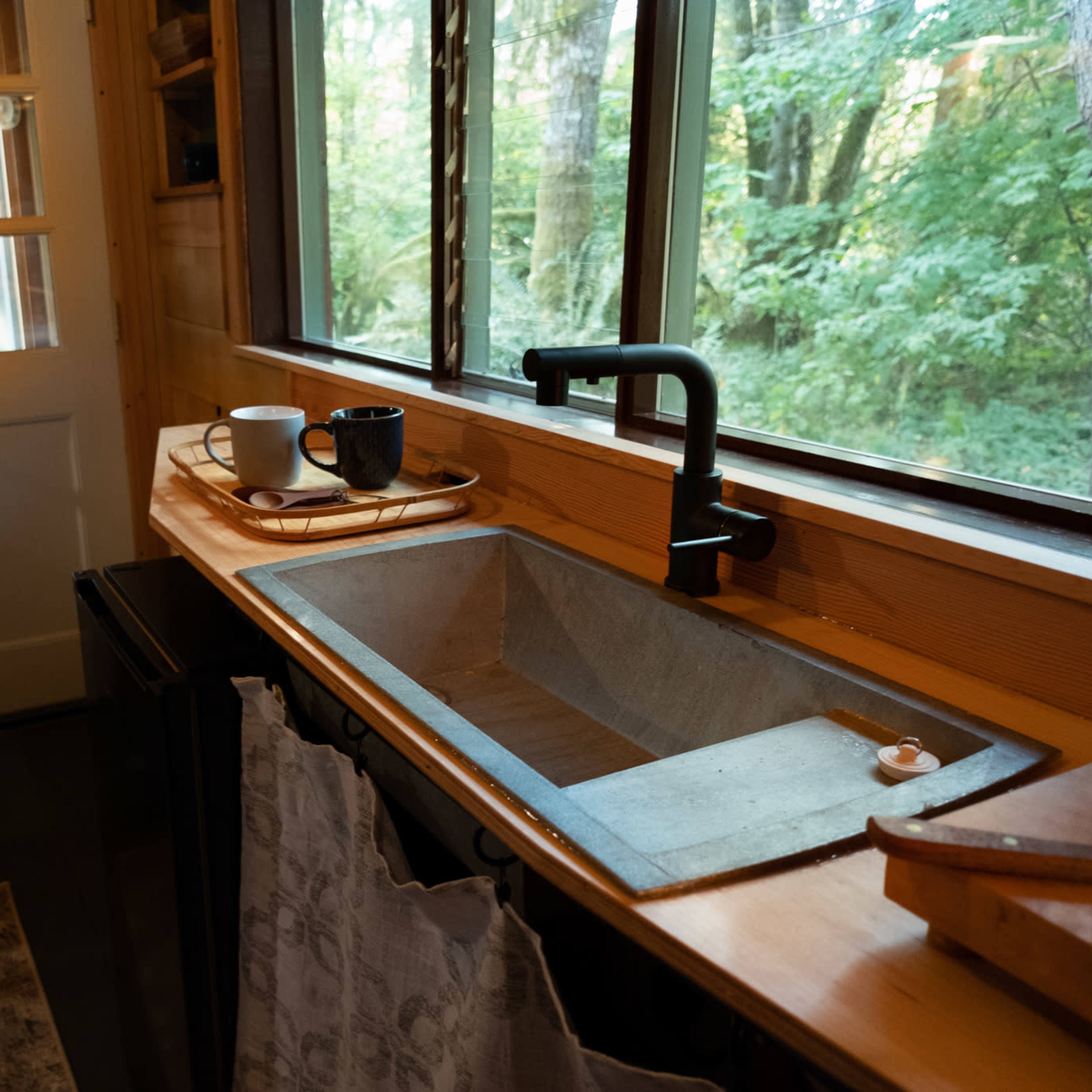 A small kitchen area with a concrete sink, wooden countertop, and large windows overlooking a green landscape.
