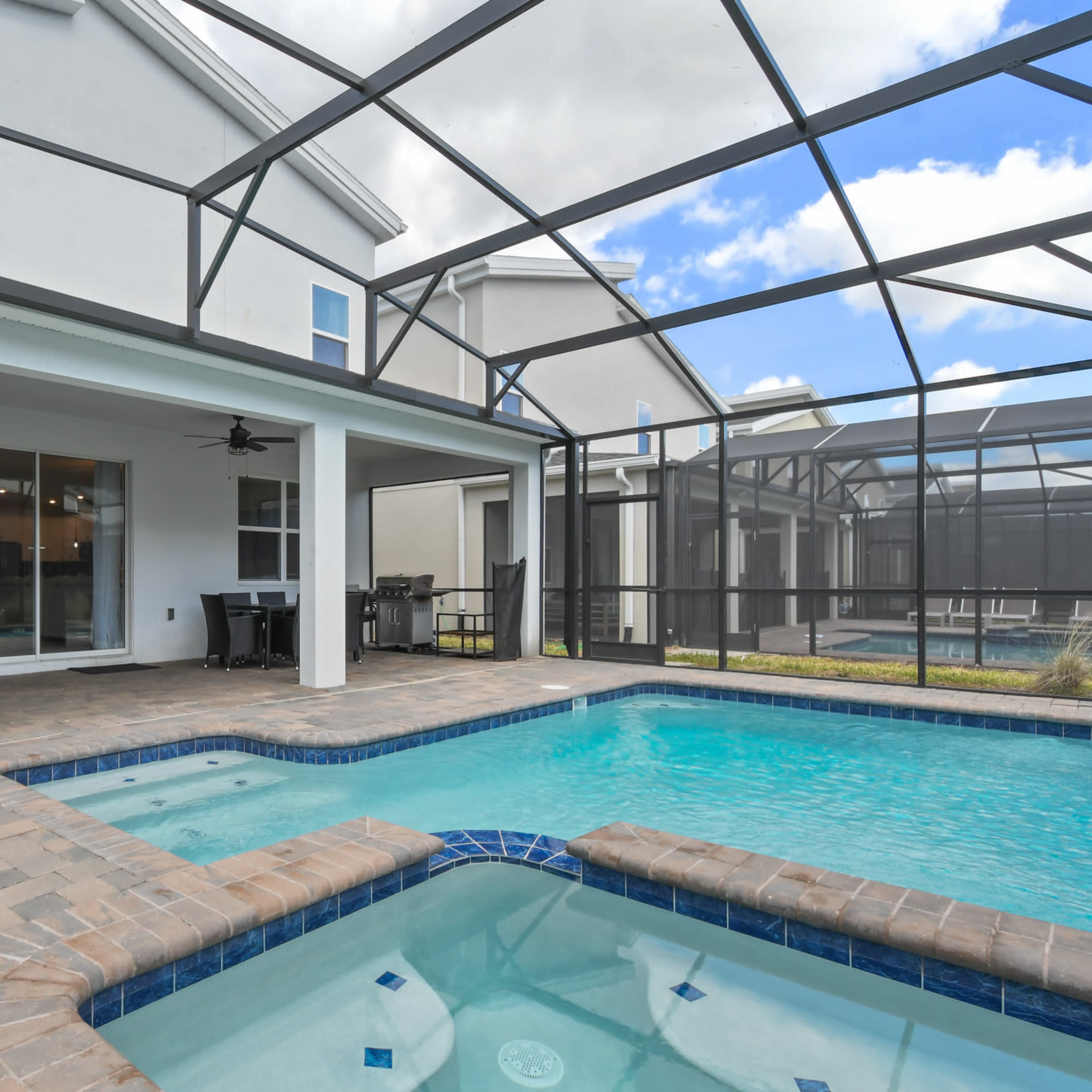 A screened-in patio area with a swimming pool and a hot tub, surrounded by outdoor furniture and a view of adjacent structures.