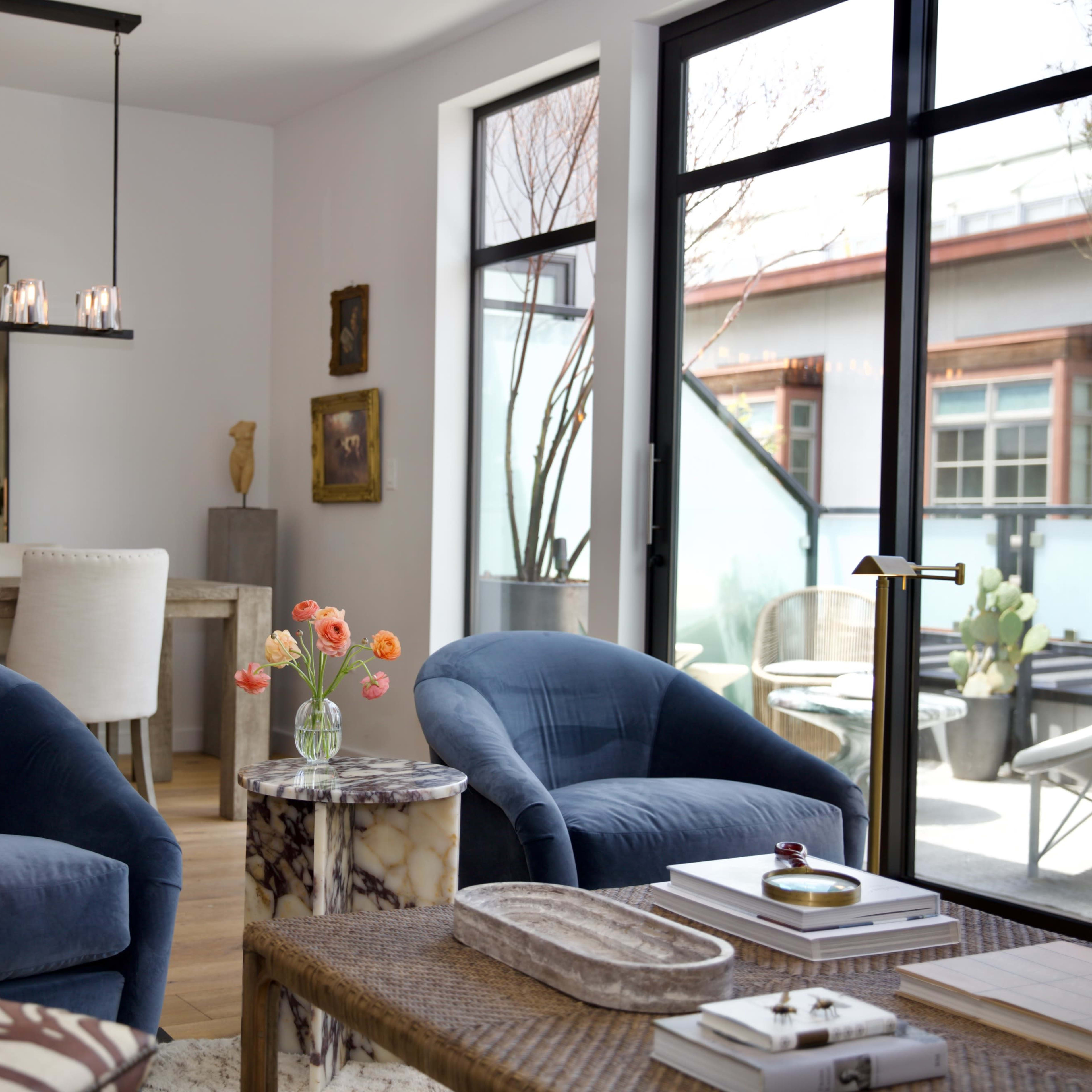 A modern living room with two blue armchairs facing a small round table, a dining area in the background, and large windows letting in natural light.