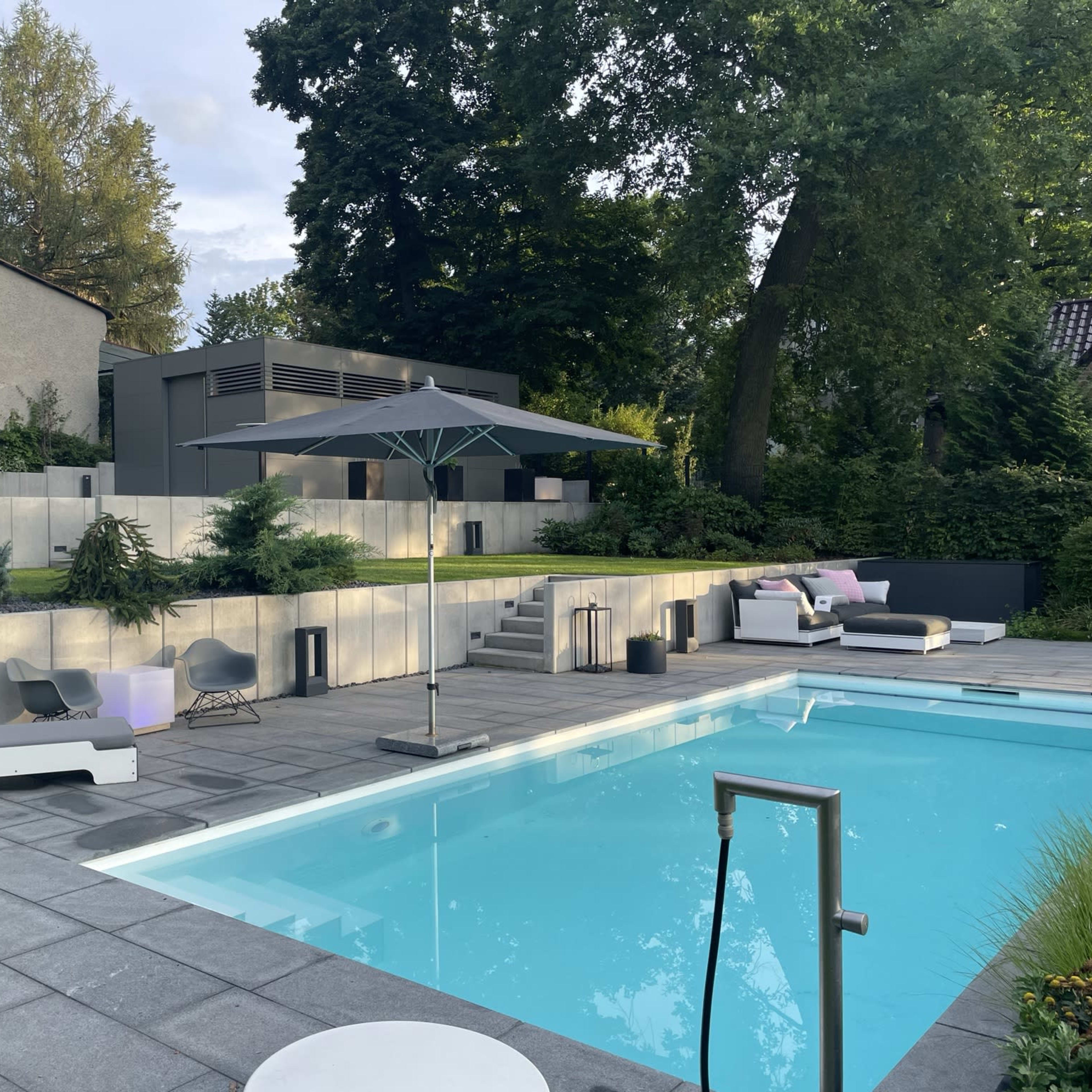 A modern backyard with a swimming pool, lounge chairs, an umbrella, and landscaped greenery surrounding a contemporary house.