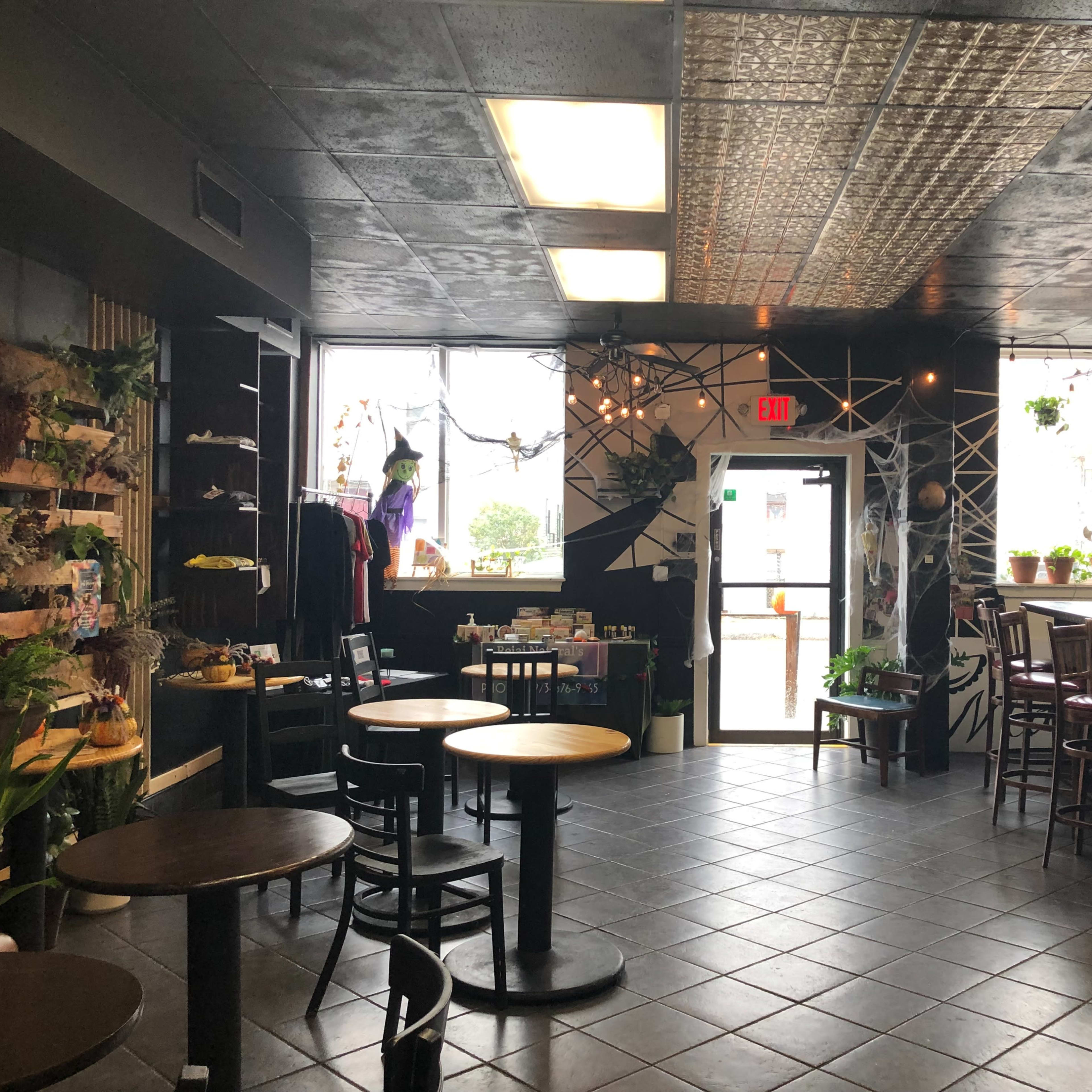 A café interior with black tables and chairs, plants on shelves, and large windows letting in natural light.