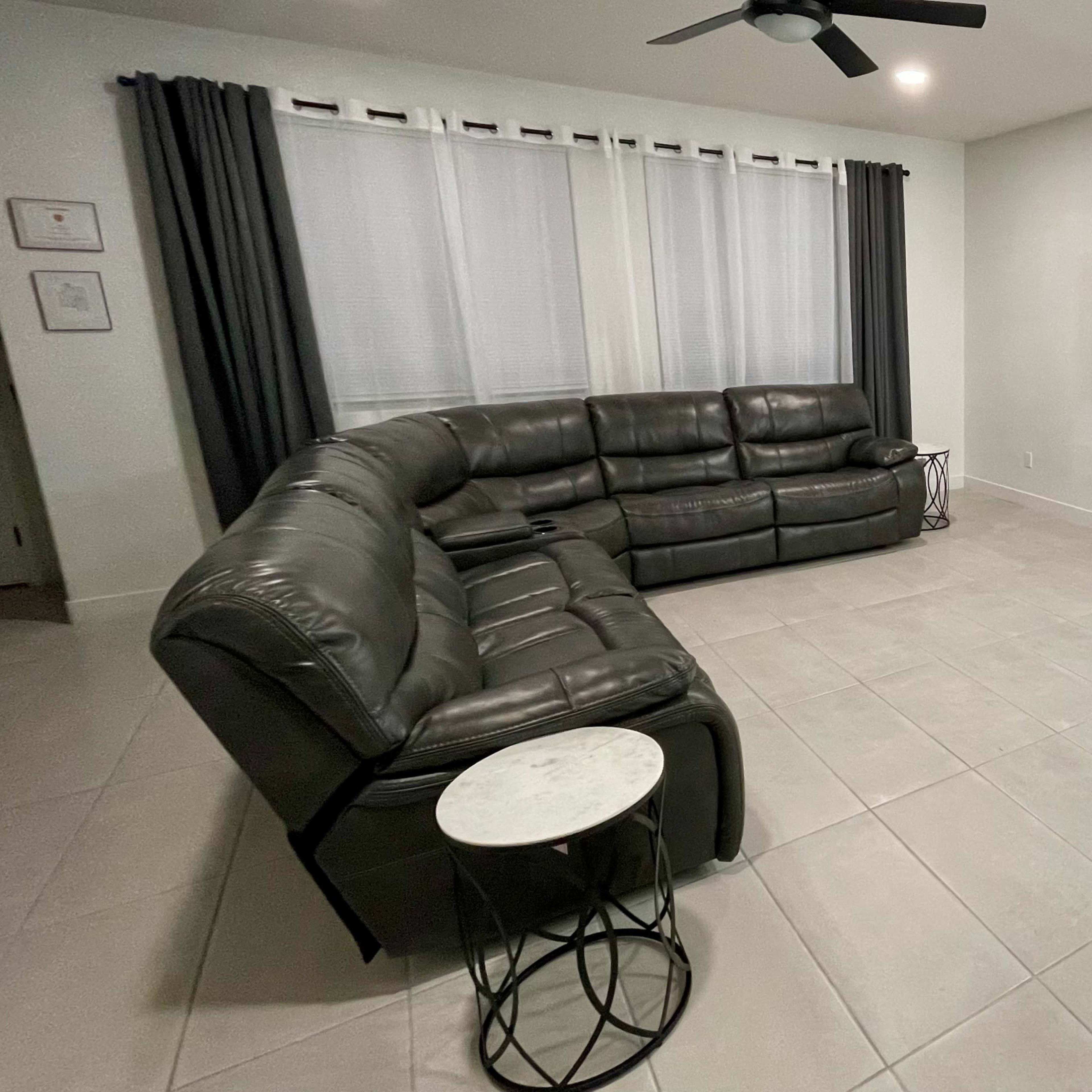 A spacious living room features a gray sectional sofa, a mounted flat-screen TV, a small black table, and a decorative lamp.