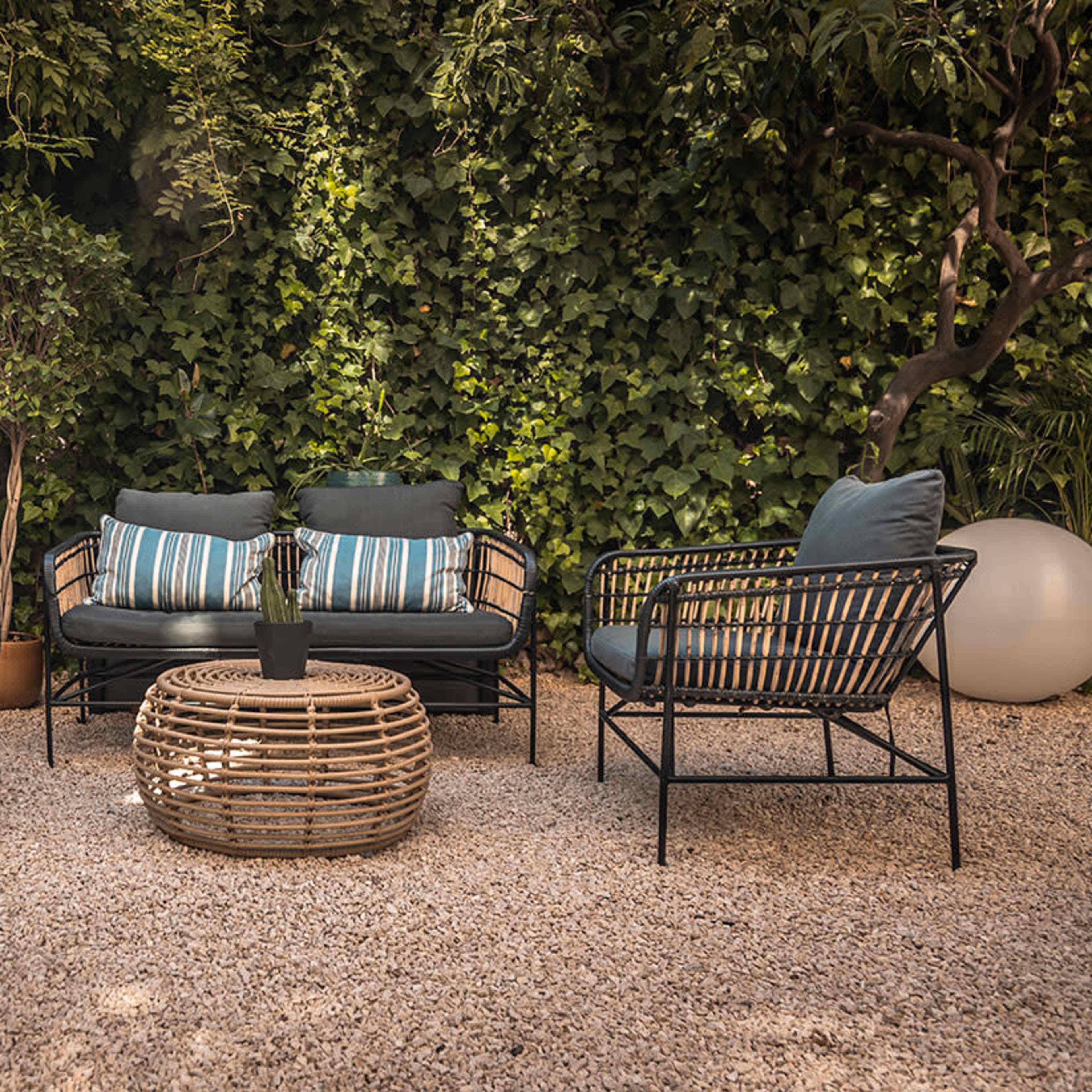 A seating area with a sofa and four chairs, surrounded by greenery and featuring a round woven coffee table on a gravel surface.