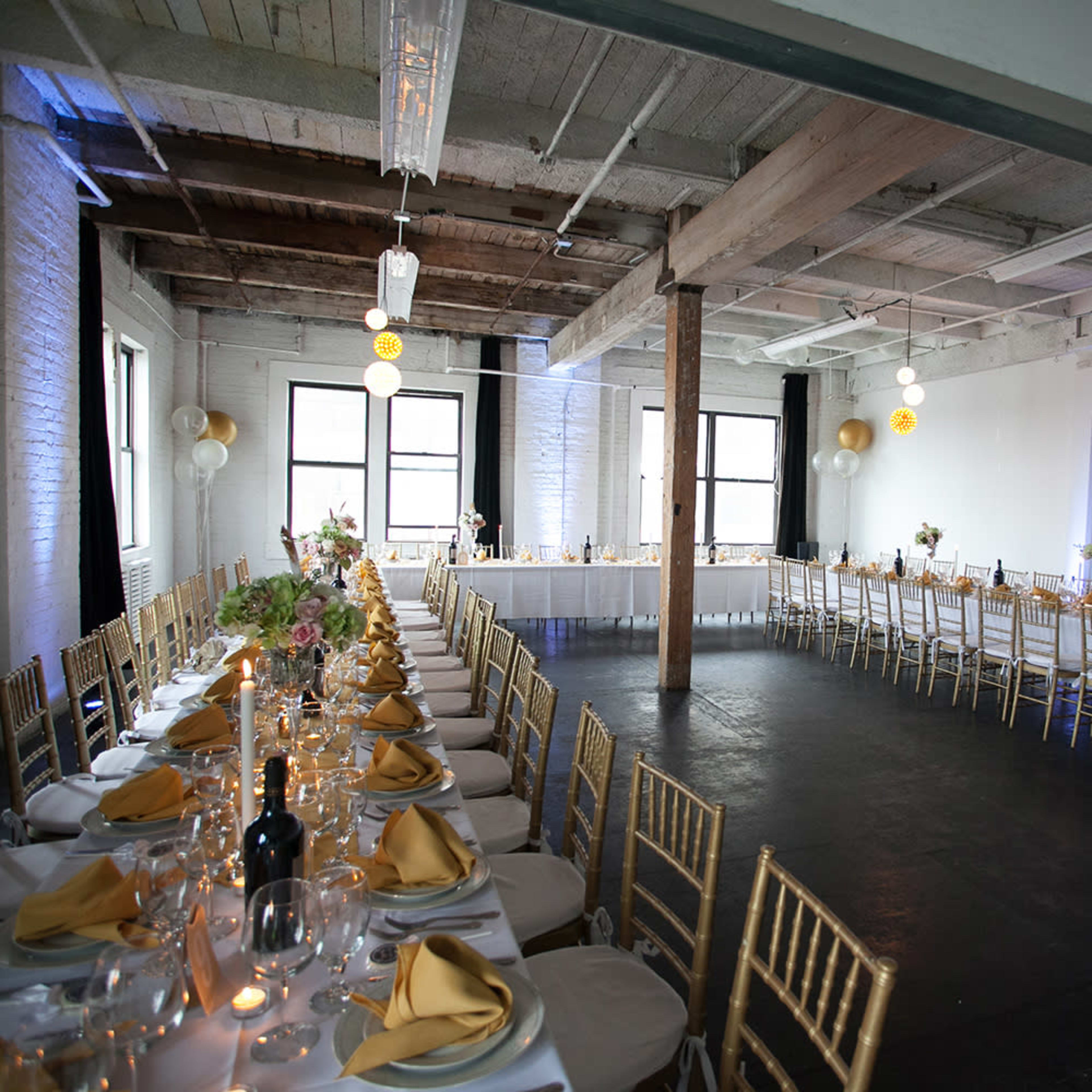 A spacious event room set up for a formal dining occasion, featuring long tables decorated with gold tableware and centerpieces.