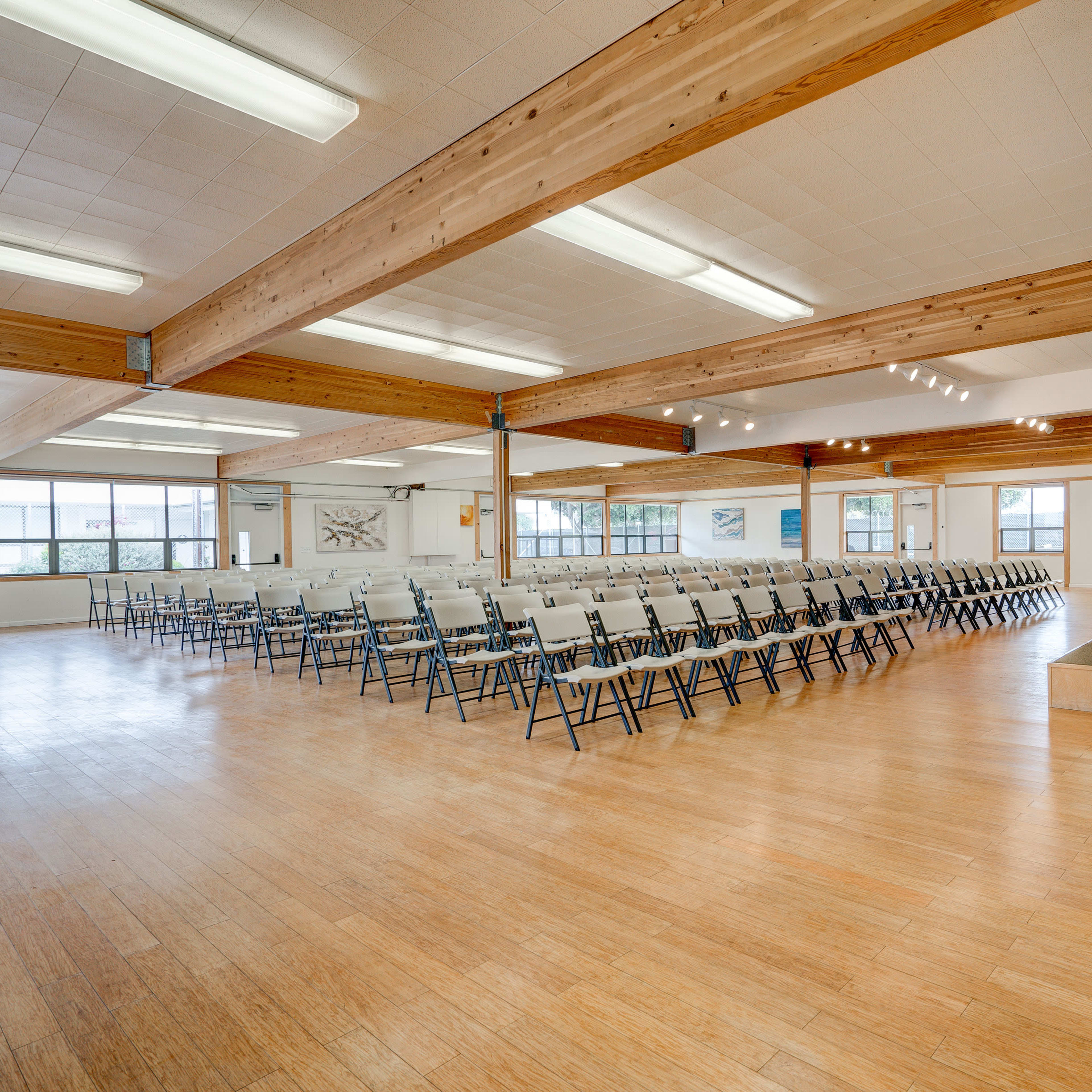 A spacious interior room features rows of chairs facing a podium on a raised platform with large windows allowing natural light.