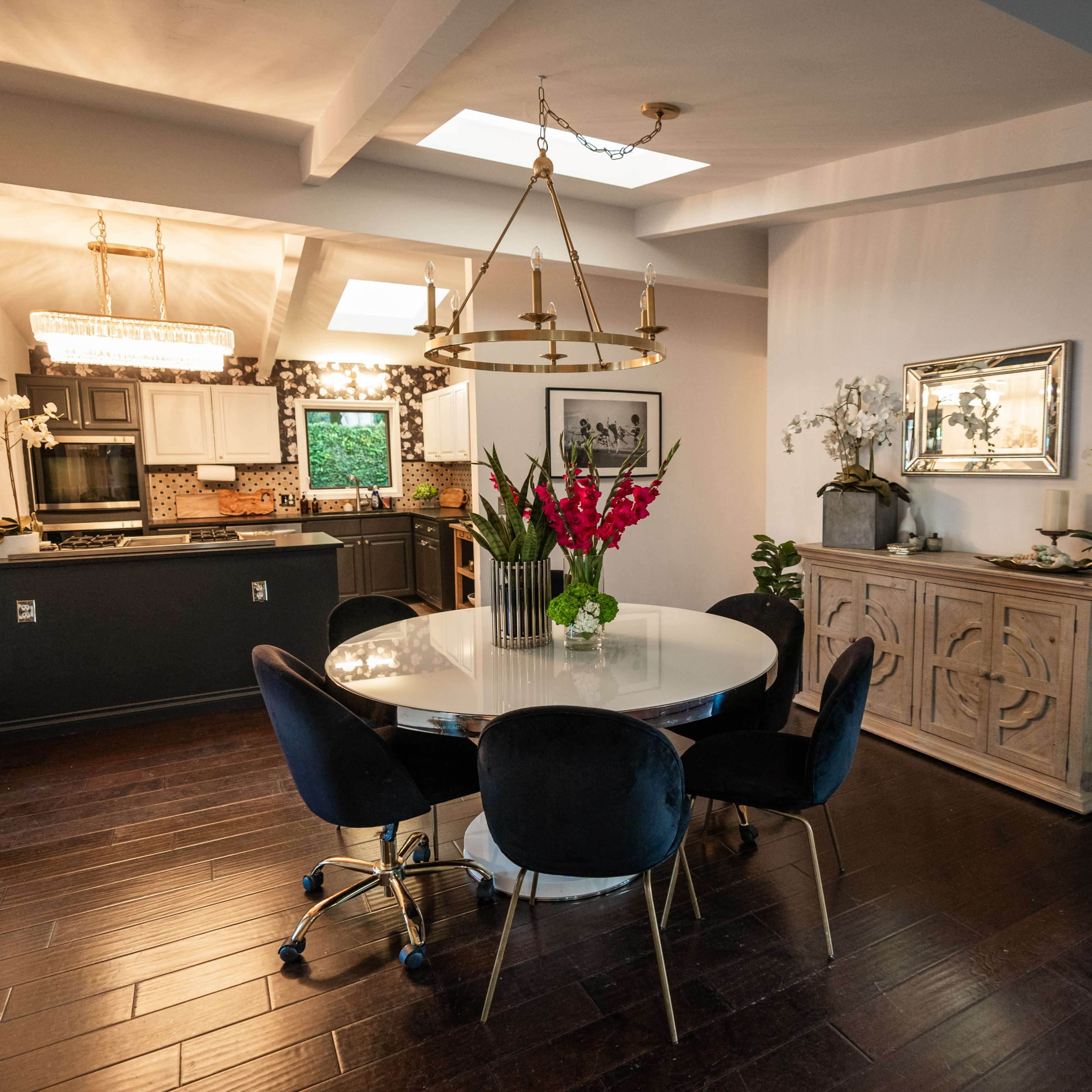 A modern dining area with a round table, four chairs, and a sideboard against the wall, surrounded by plants and featuring a kitchen in the background.
