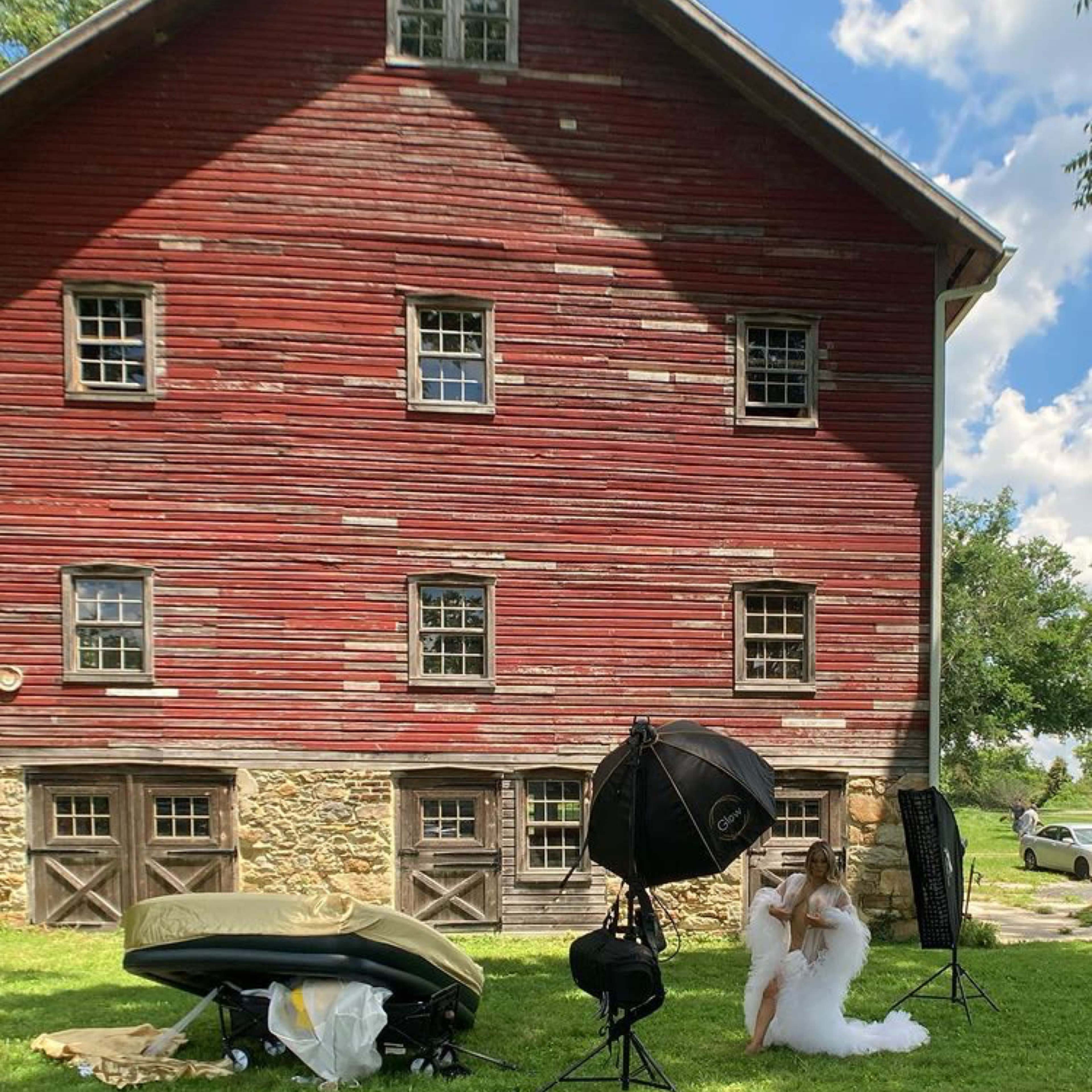 A woman in a white dress poses near photography equipment in front of a red barn with multiple windows.