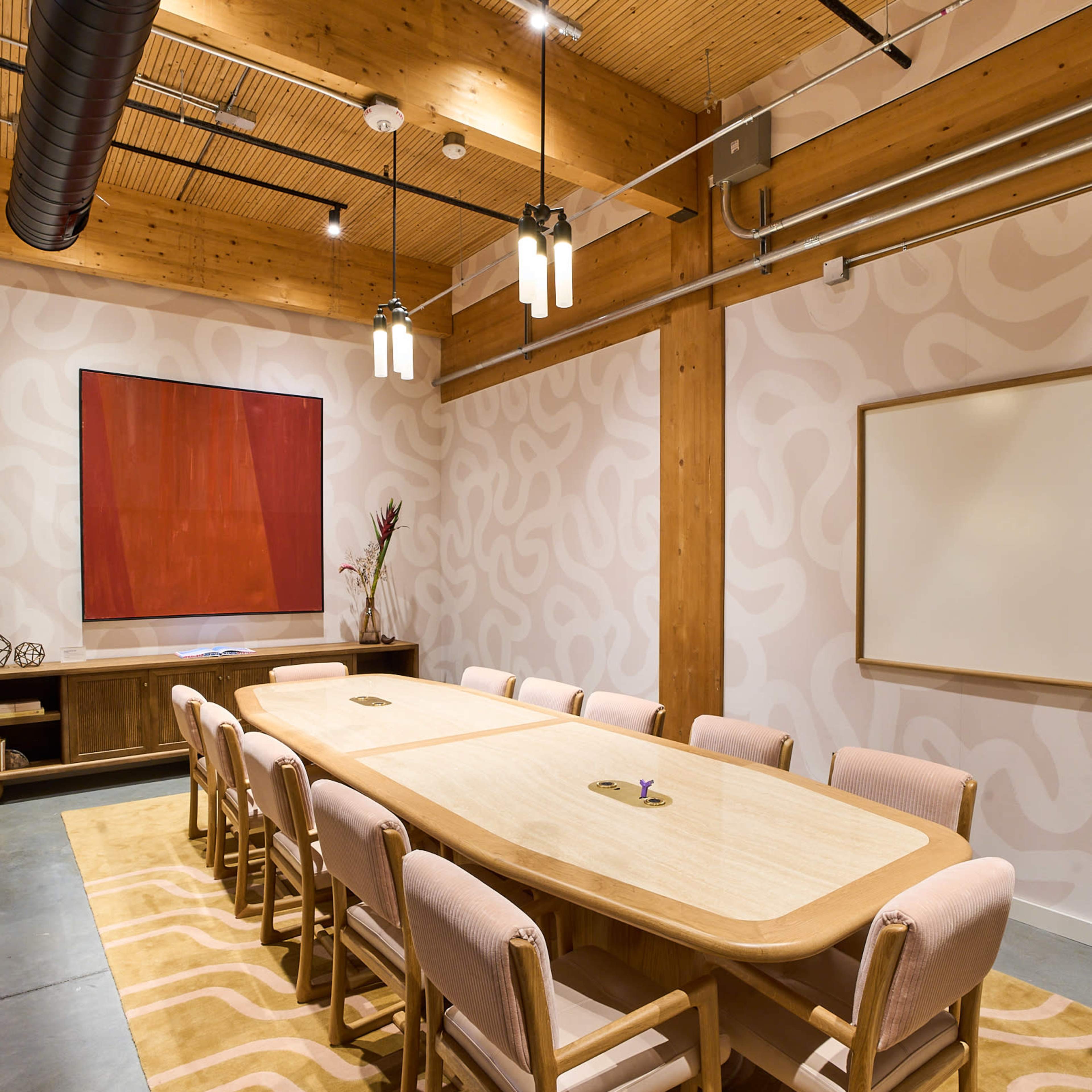 A modern conference room featuring a large wooden table surrounded by twelve chairs on a patterned rug, with minimalistic decor including a wall art piece and a whiteboard.