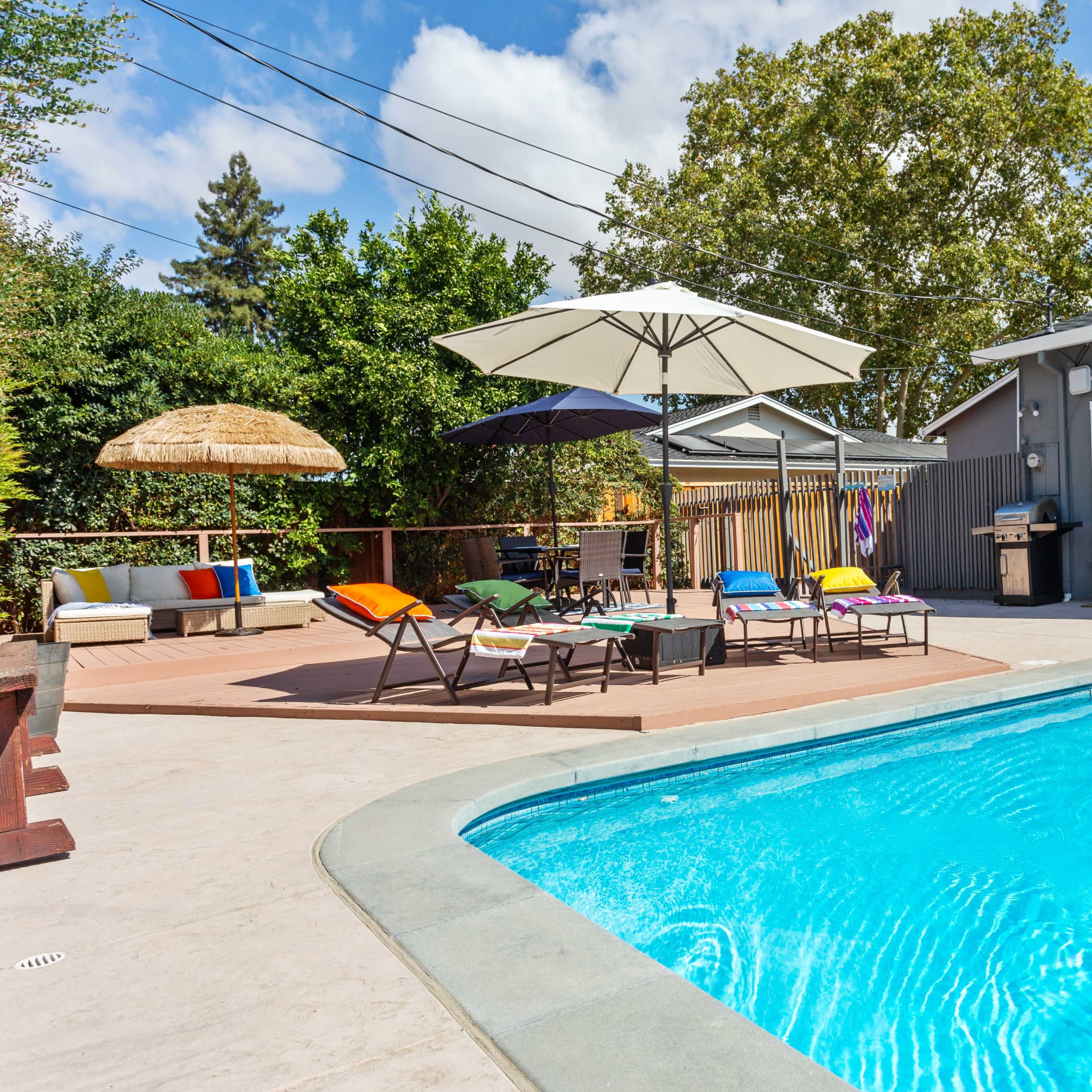 A backyard featuring a swimming pool surrounded by lounge chairs, a large umbrella, and outdoor seating areas.