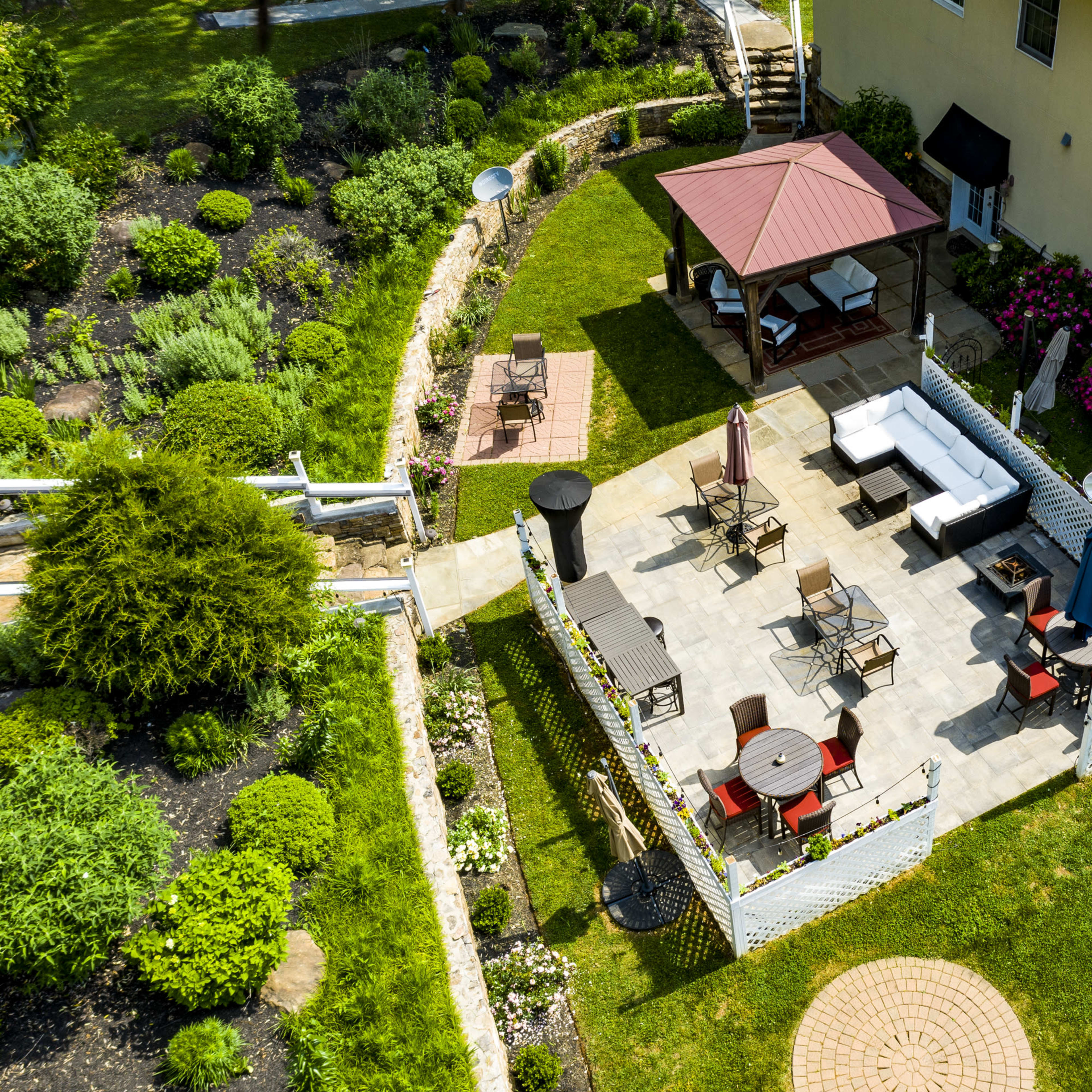 An aerial view of a landscaped backyard featuring a paved patio with seating arrangements, a gazebo, and garden plants.