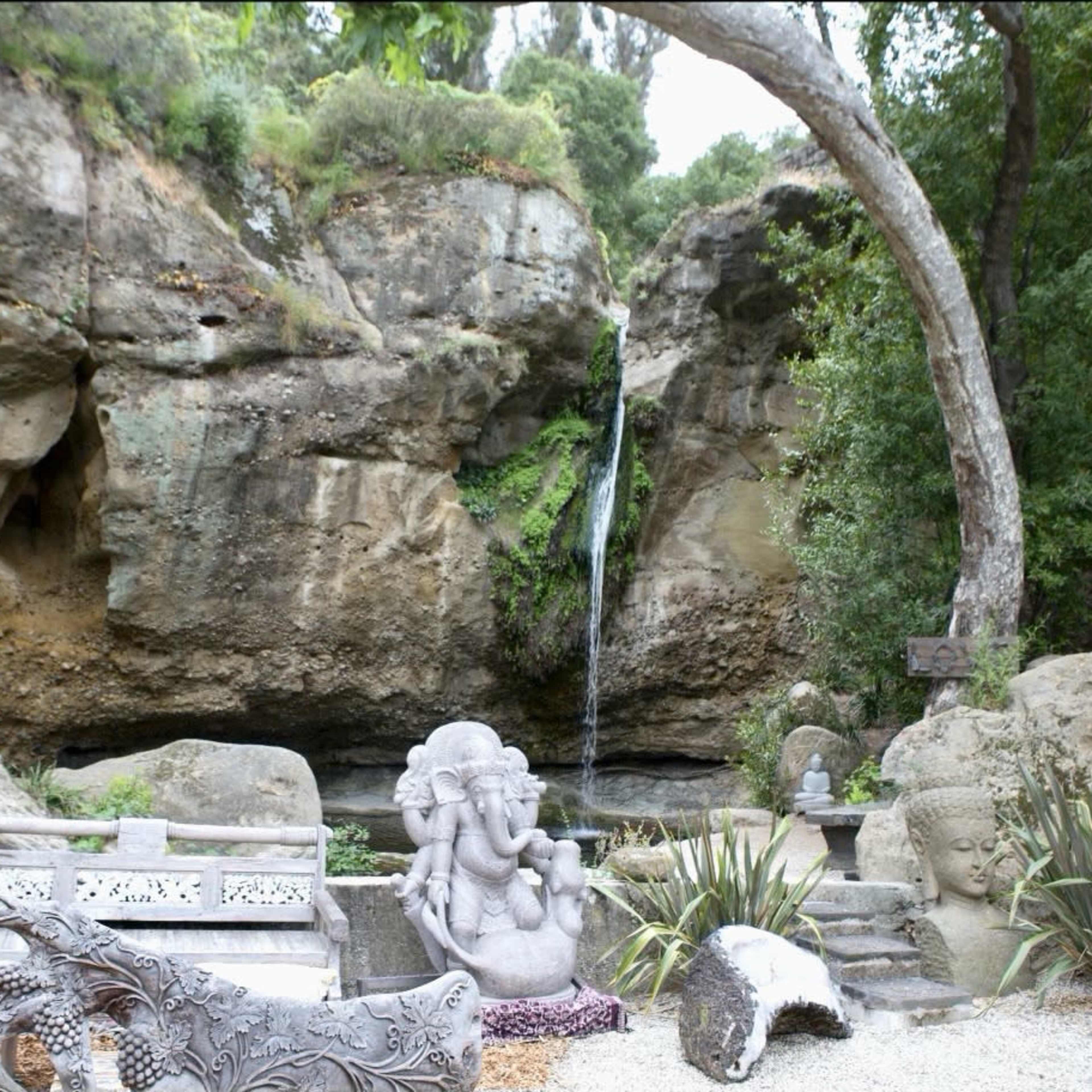 A serene outdoor scene with stone sculptures, a small waterfall cascading from a rocky cliff, and lush greenery surrounding the area.